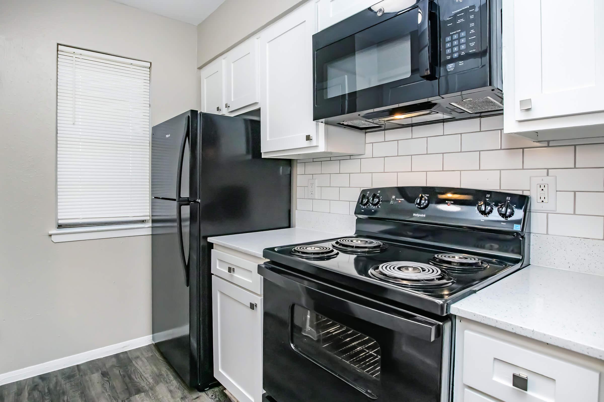A modern kitchen featuring a black refrigerator, a black electric stove with four burners, and a built-in microwave above the stove. White cabinets and a light-colored countertop complement the black appliances. A window with blinds provides natural light. The flooring is a dark wood finish.