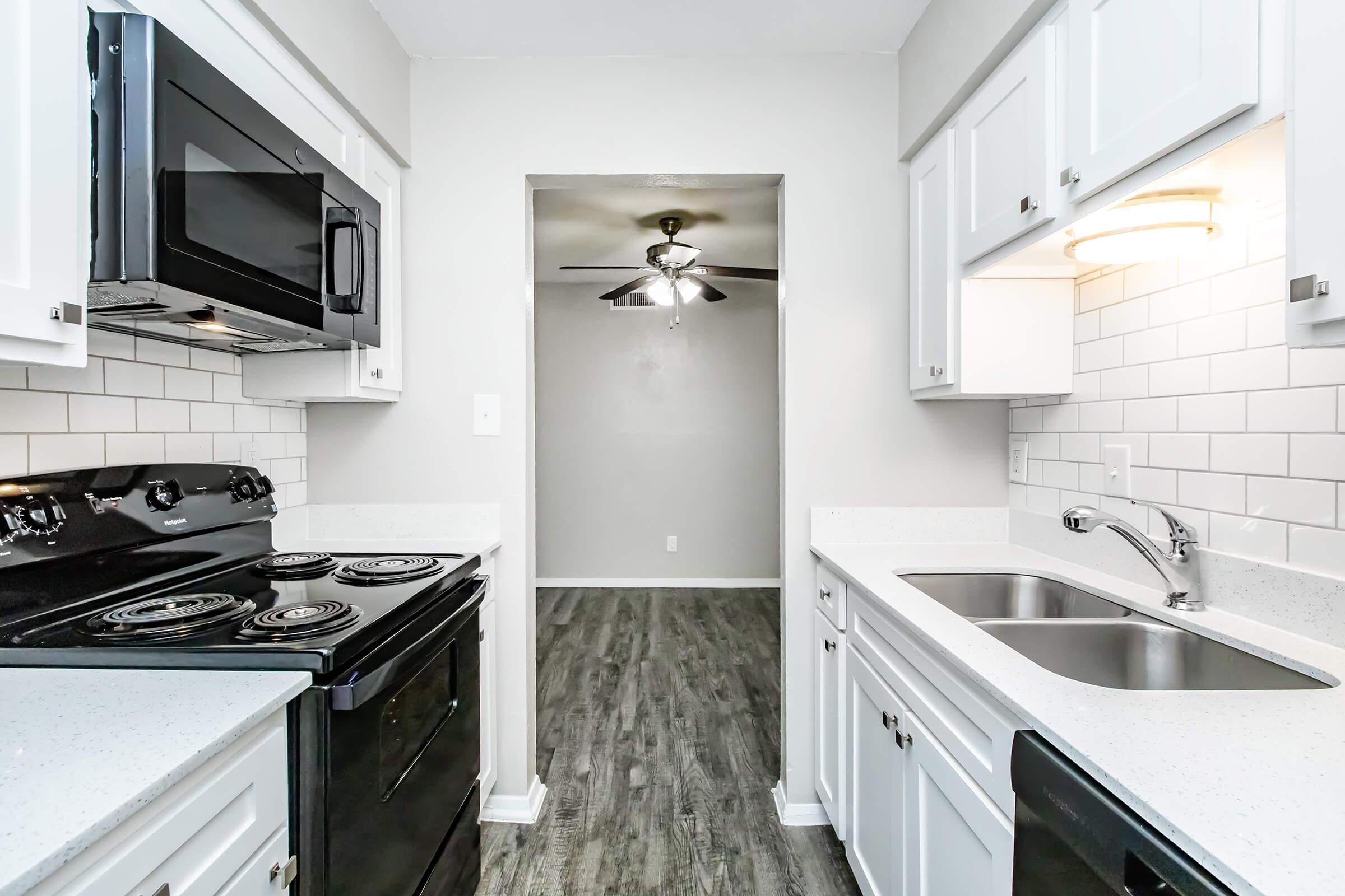 Modern kitchen featuring a black stove, microwave, and stainless steel sink. White cabinetry with a clean subway tile backsplash. Light wood-like flooring enhances the bright and spacious feel, leading to a doorway that opens into a well-lit living area.
