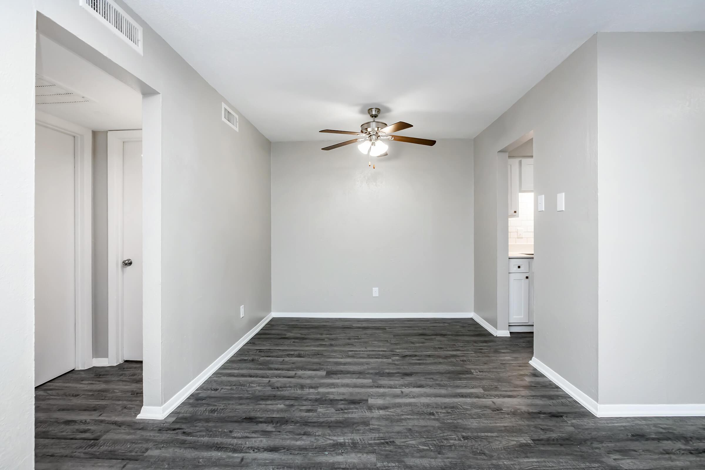 Spacious living area with gray walls, a ceiling fan, and laminate flooring. Two doorways lead to adjacent rooms, one of which appears to connect to a kitchen. The space is well-lit, creating a welcoming atmosphere.