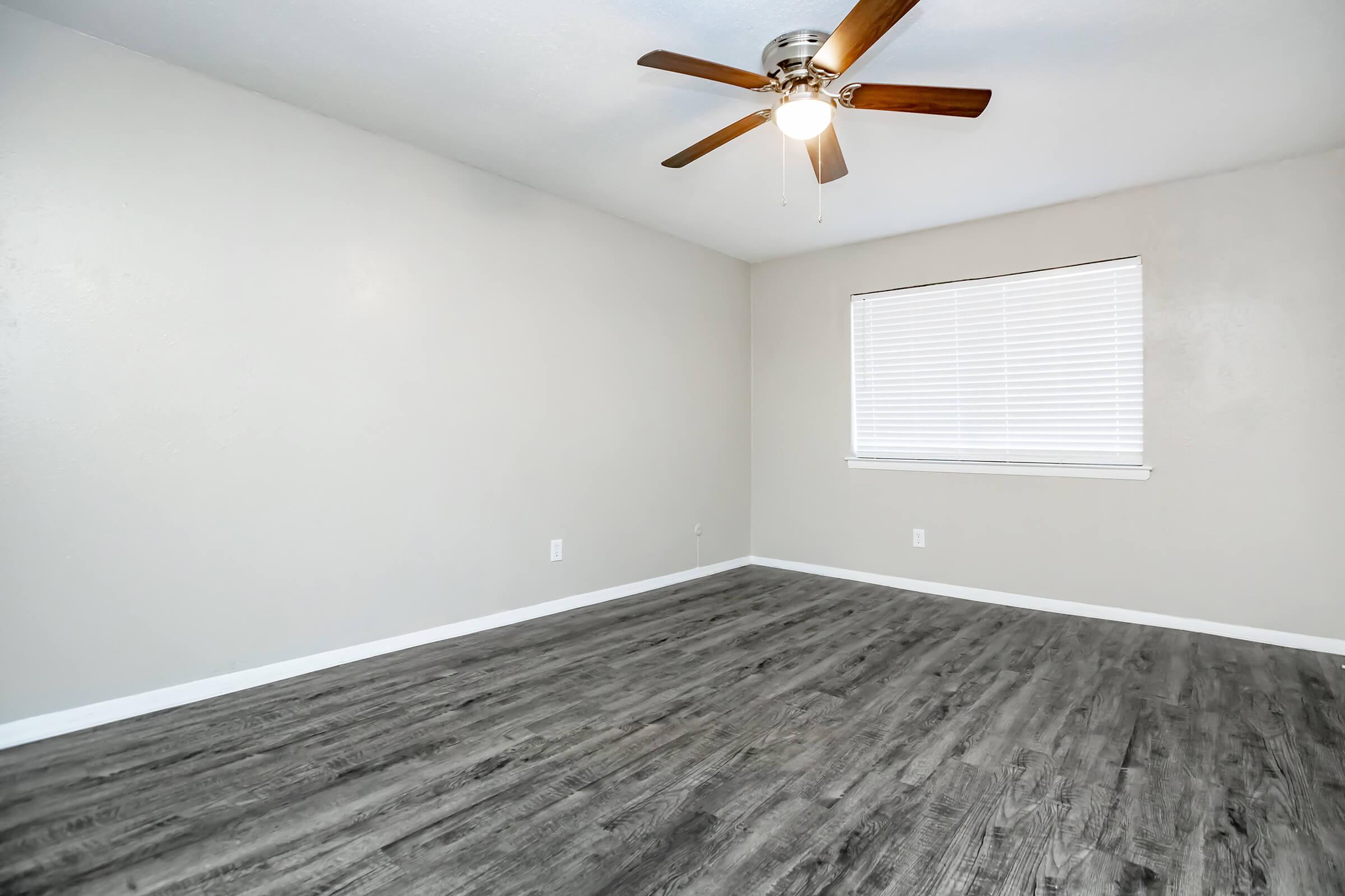 Empty room with light gray walls and a ceiling fan featuring wooden blades. A large window with white blinds allows natural light to enter, and the floor is covered with dark wood-like laminate. The room's design is simple and modern, ideal for personalization.