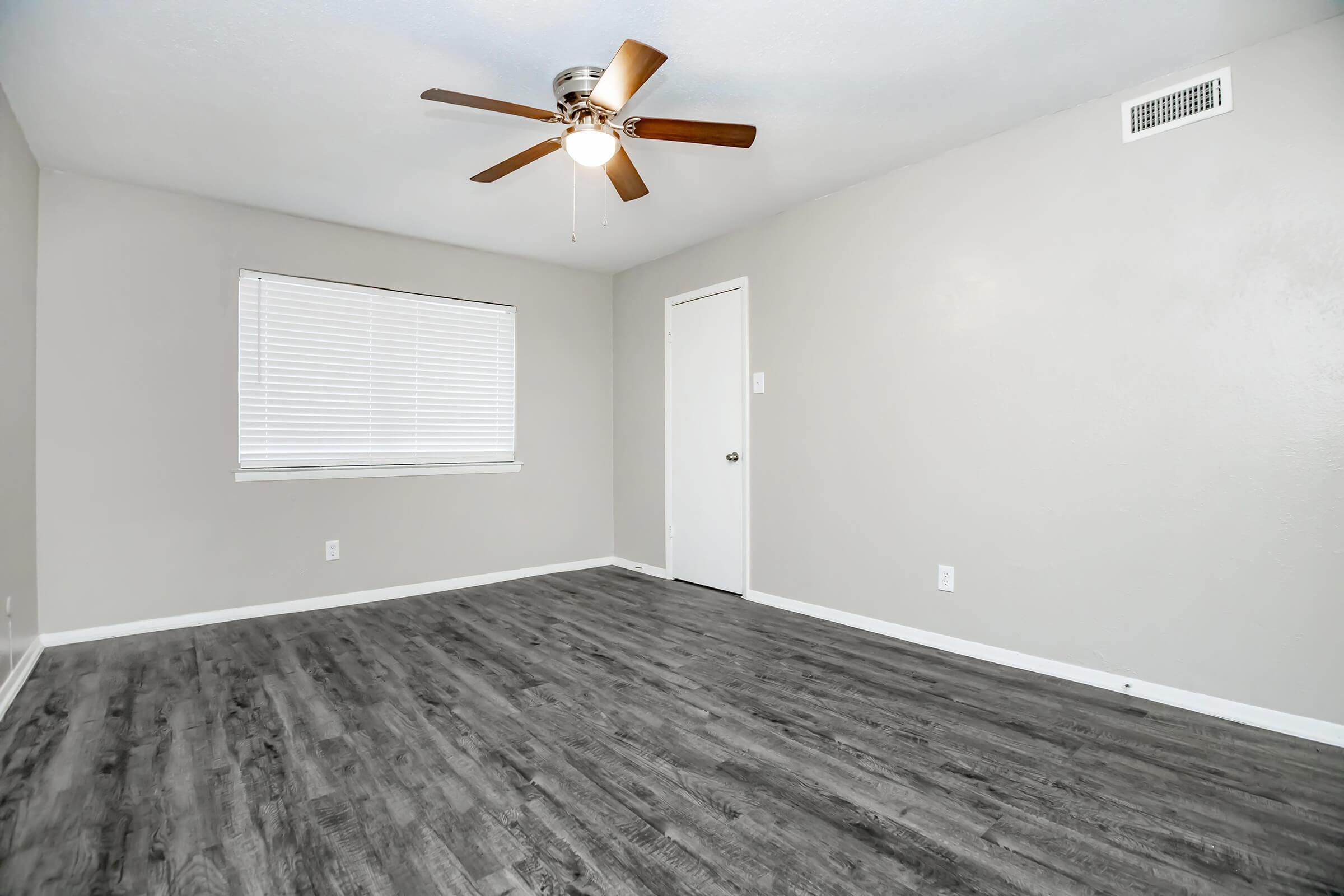 A spacious, empty room featuring light gray walls and a modern ceiling fan. The flooring is dark wood laminate, and there is a window with white blinds allowing natural light. A plain white door is visible on one side, enhancing the room's minimalist aesthetic.