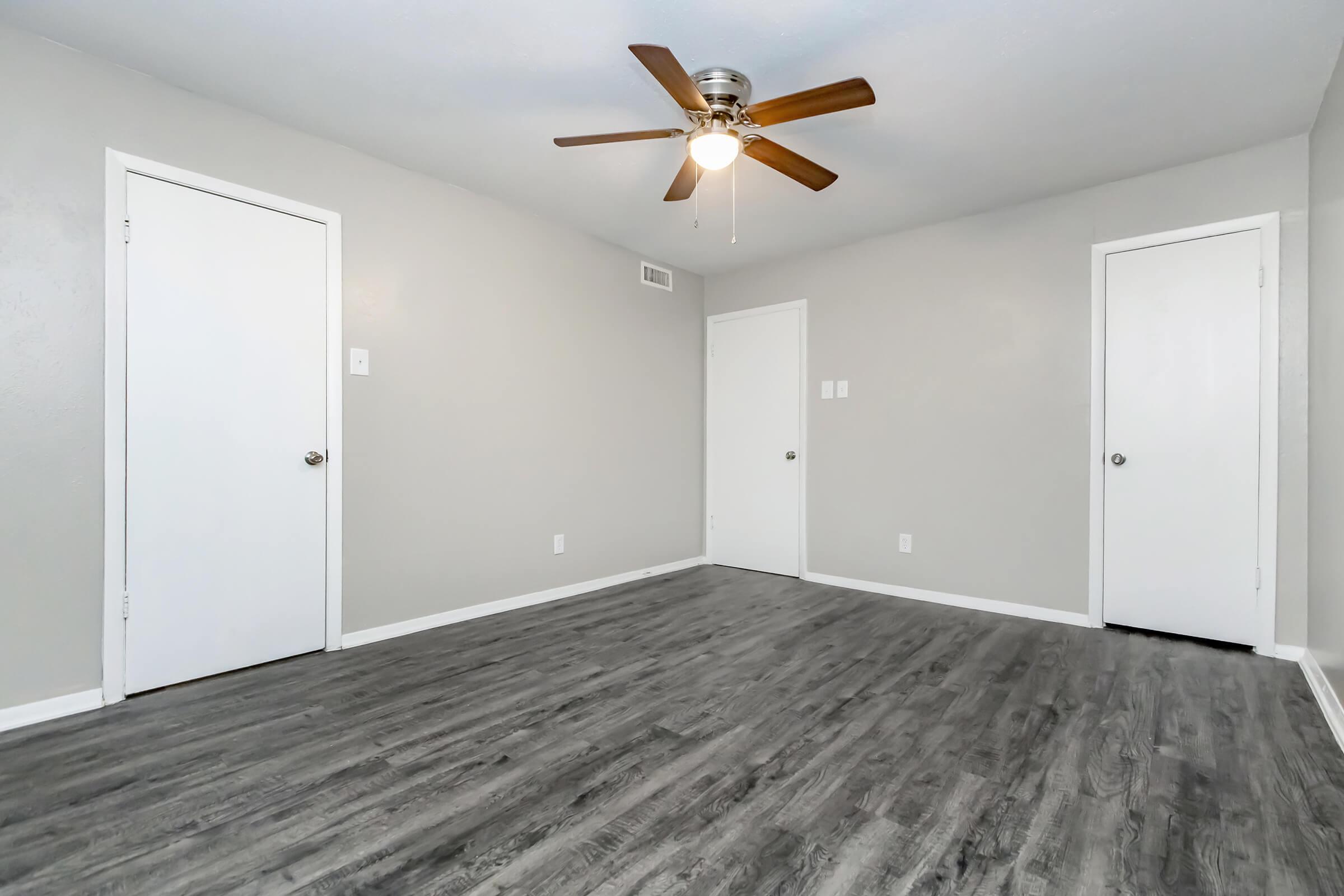A spacious, empty room featuring a light gray wall color, two closed white doors, and a ceiling fan with wooden blades. The floor is covered with dark wood-like laminate, creating a modern and clean aesthetic. Natural light may be coming in from an unseen window.