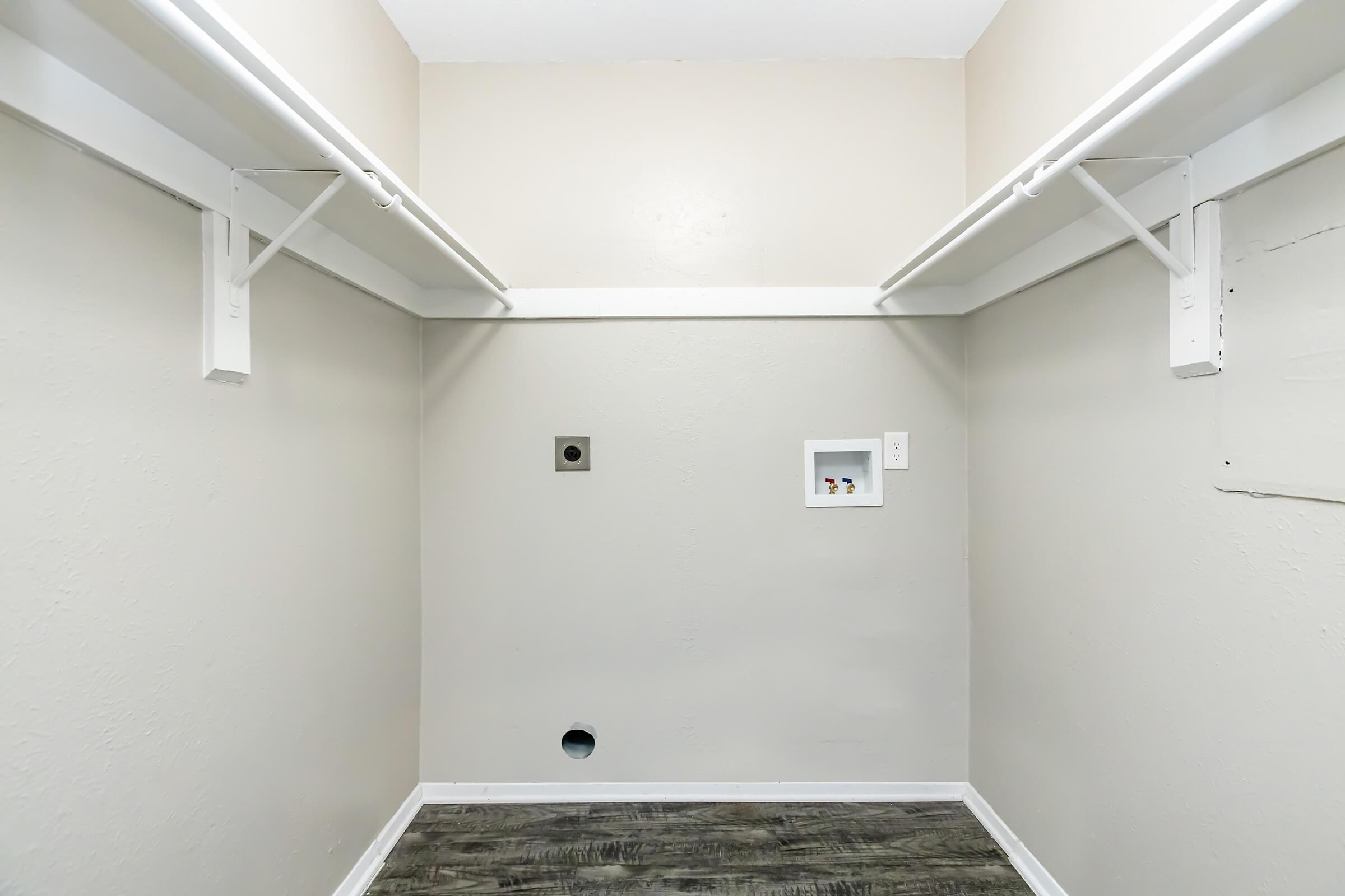Empty laundry room with light beige walls, two white overhead shelves, and a dark gray floor. A wall outlet and a connection point are visible on the right wall, while a hole in the floor suggests plumbing access. The space is well-lit and clear of clutter.