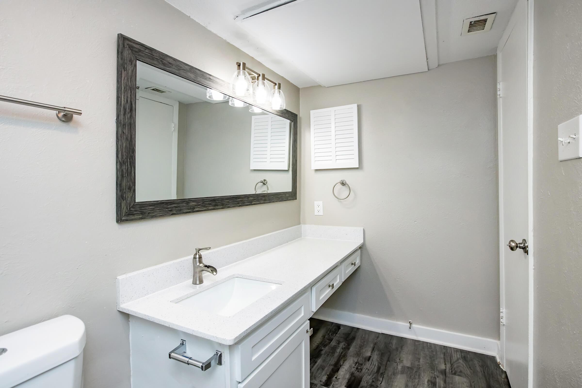 Modern bathroom interior featuring a marble countertop with a single sink, a large mirror above, and contemporary lighting fixtures. Walls are painted neutral colors, and there is a towel ring next to the sink. A white toilet is visible on the left, with a door leading to another area of the space.