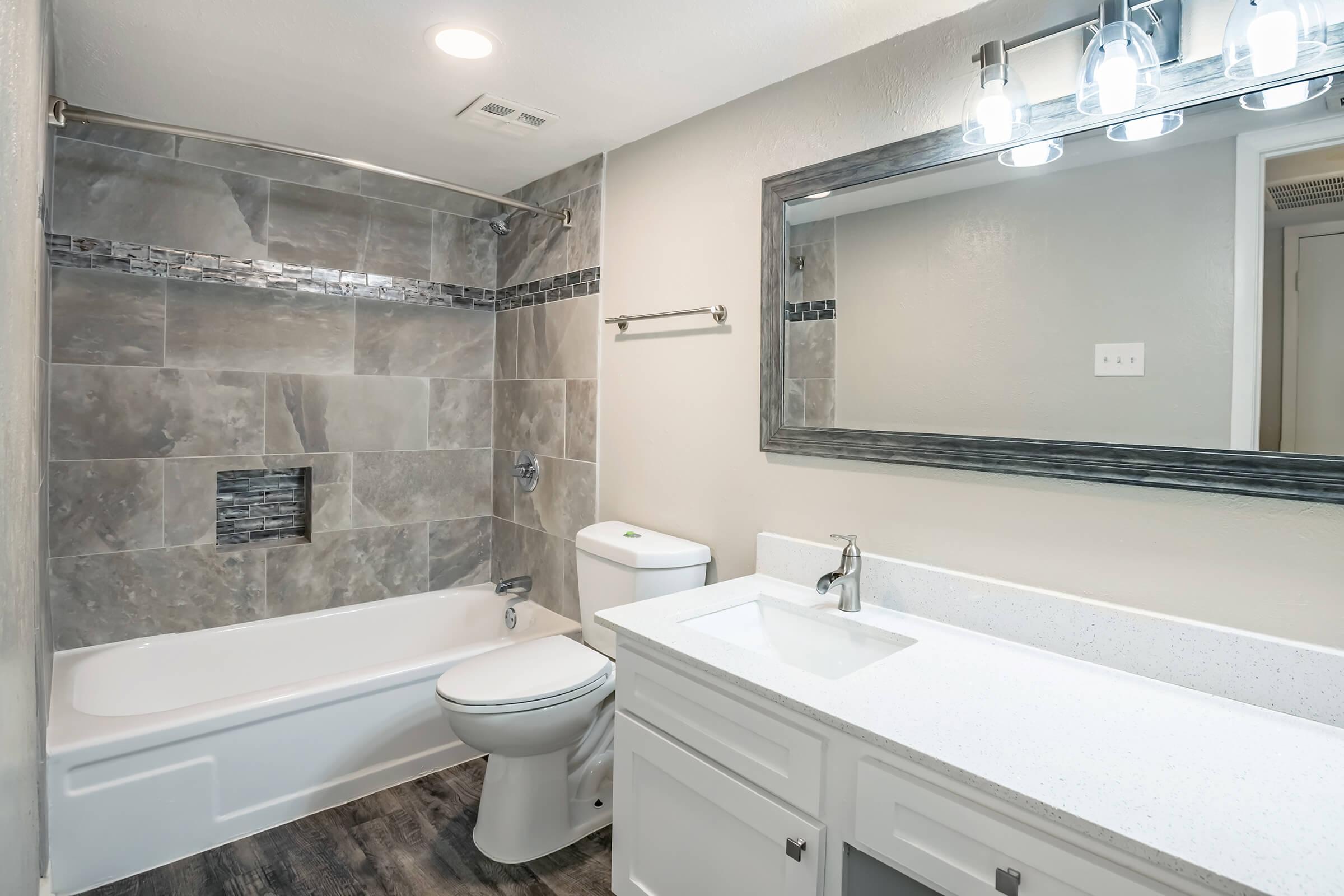A modern bathroom featuring a bathtub with a shower, grey tiled walls, a large mirror above a white vanity with a sink, and a toilet. There is a sleek light fixture above the mirror, and the flooring is dark wood laminate. The overall color scheme is neutral and contemporary.
