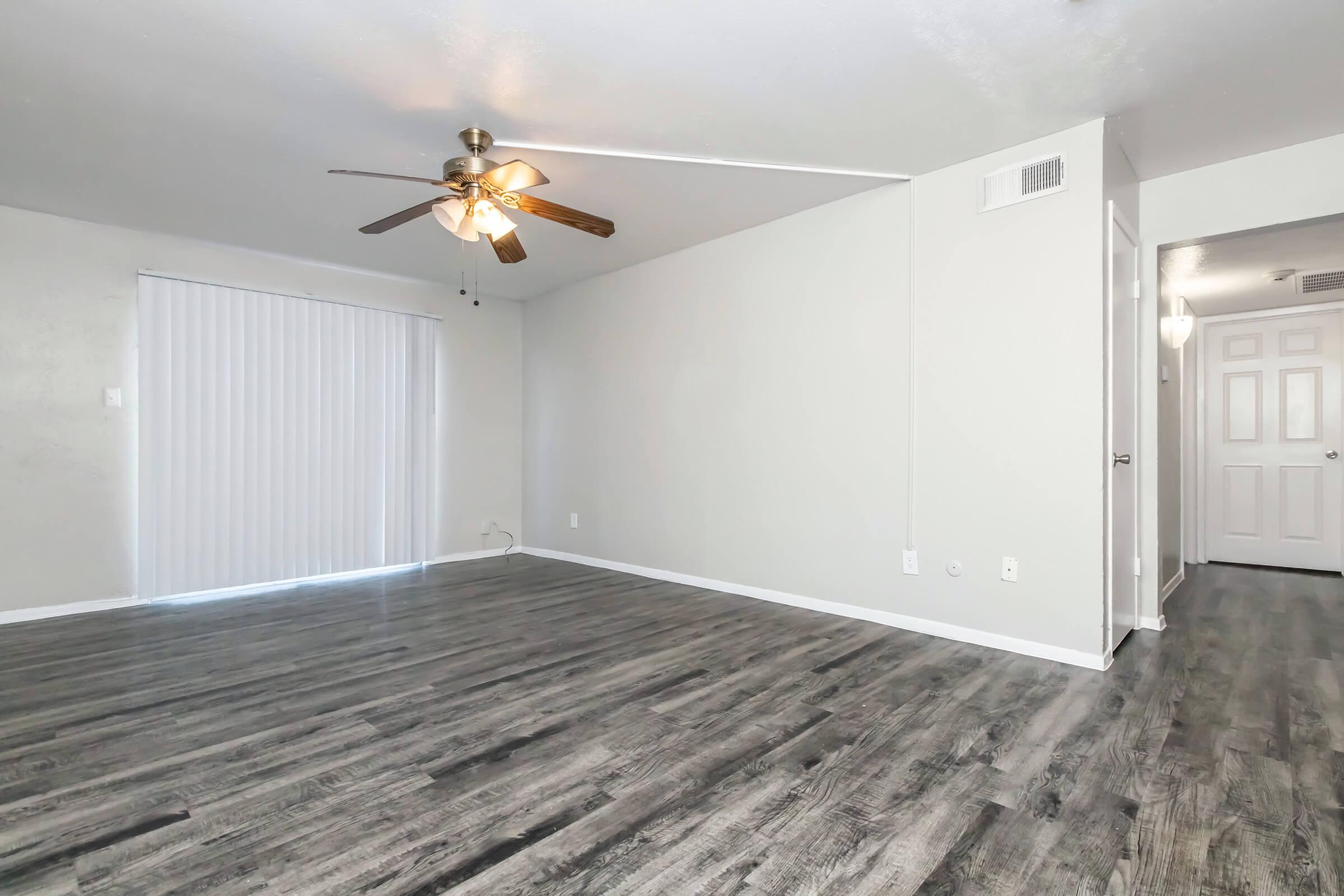 Spacious empty living room featuring a ceiling fan, large window with vertical blinds, and light hardwood flooring. The walls are painted in a soft gray color, and a hallway leading to other rooms is visible in the background.