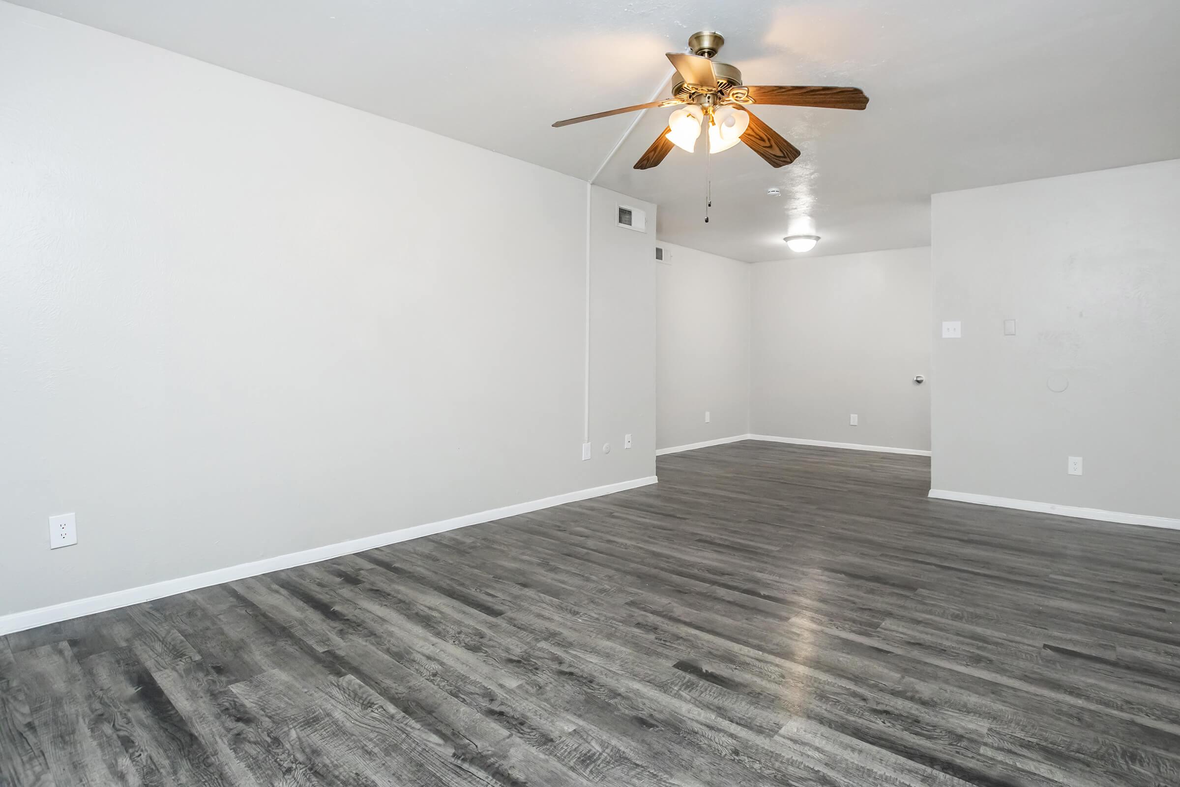 A spacious, empty room with light gray walls and dark wood-like flooring. There is a ceiling fan with wooden blades and a light fixture visible. The room has minimal furniture and is well-lit, creating a bright and open atmosphere.