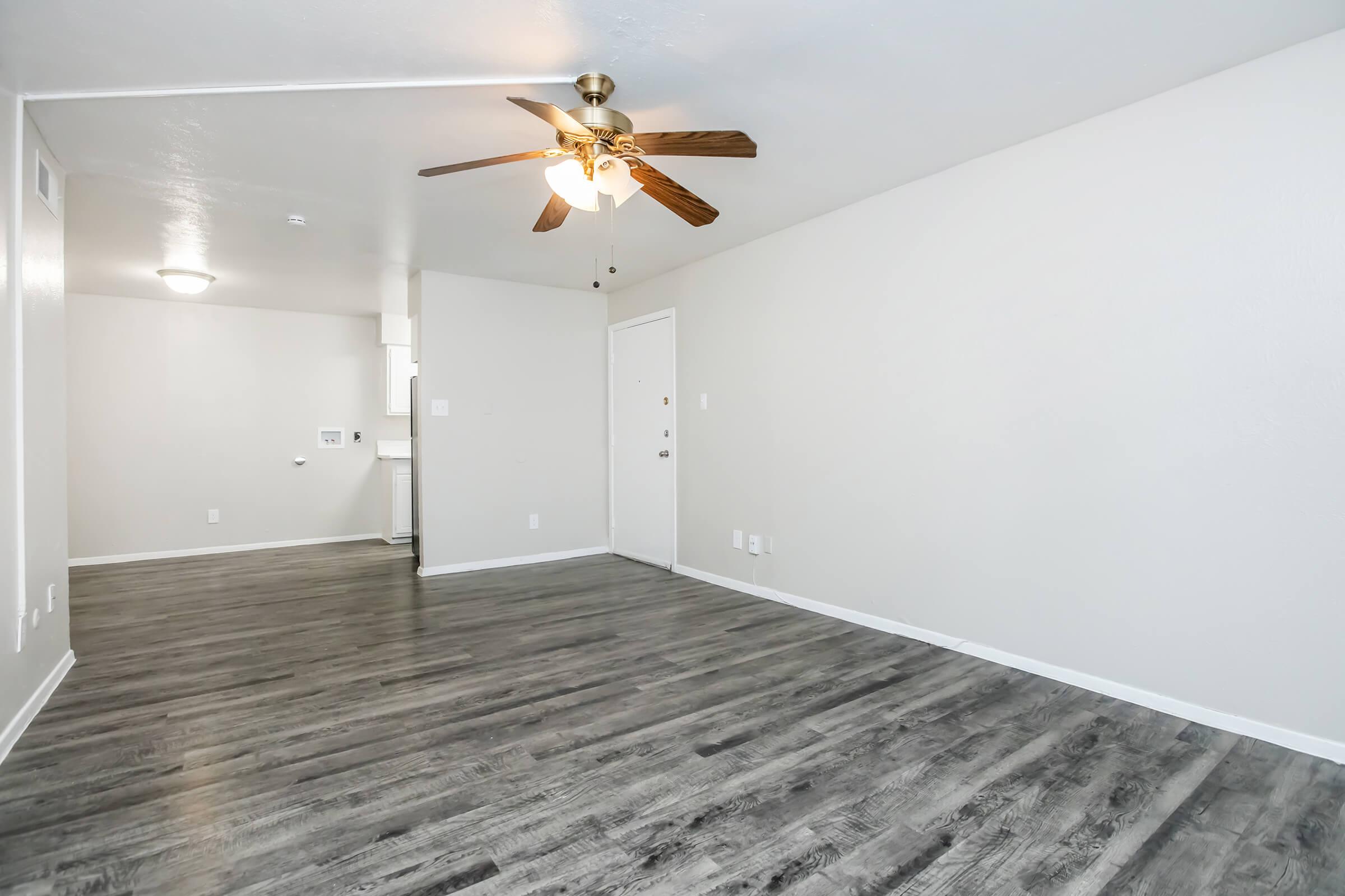 Empty room with light gray walls and wood-like flooring, featuring a ceiling fan with wooden blades. A small light fixture is visible in the ceiling, and a door leads outside. A wall-mounted outlet is located near the door, with an open space leading to another room or kitchen area in the background.