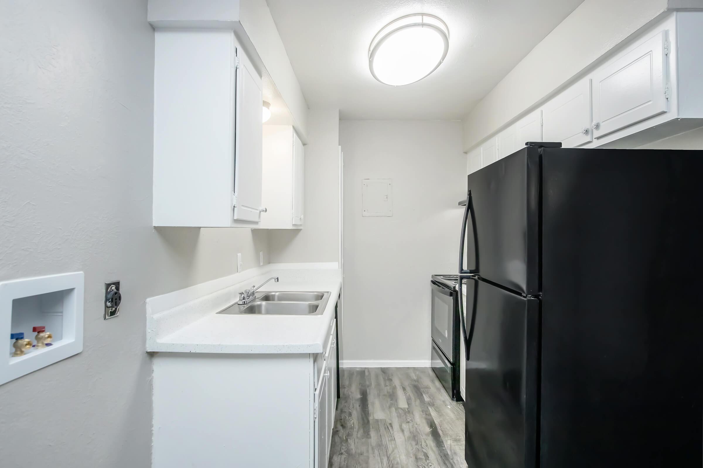 A modern kitchen featuring white cabinets, a black refrigerator, and a stainless steel stove. The countertops are white, and the floor is covered in plank-style vinyl. A circular ceiling light illuminates the space, creating a bright and clean appearance.