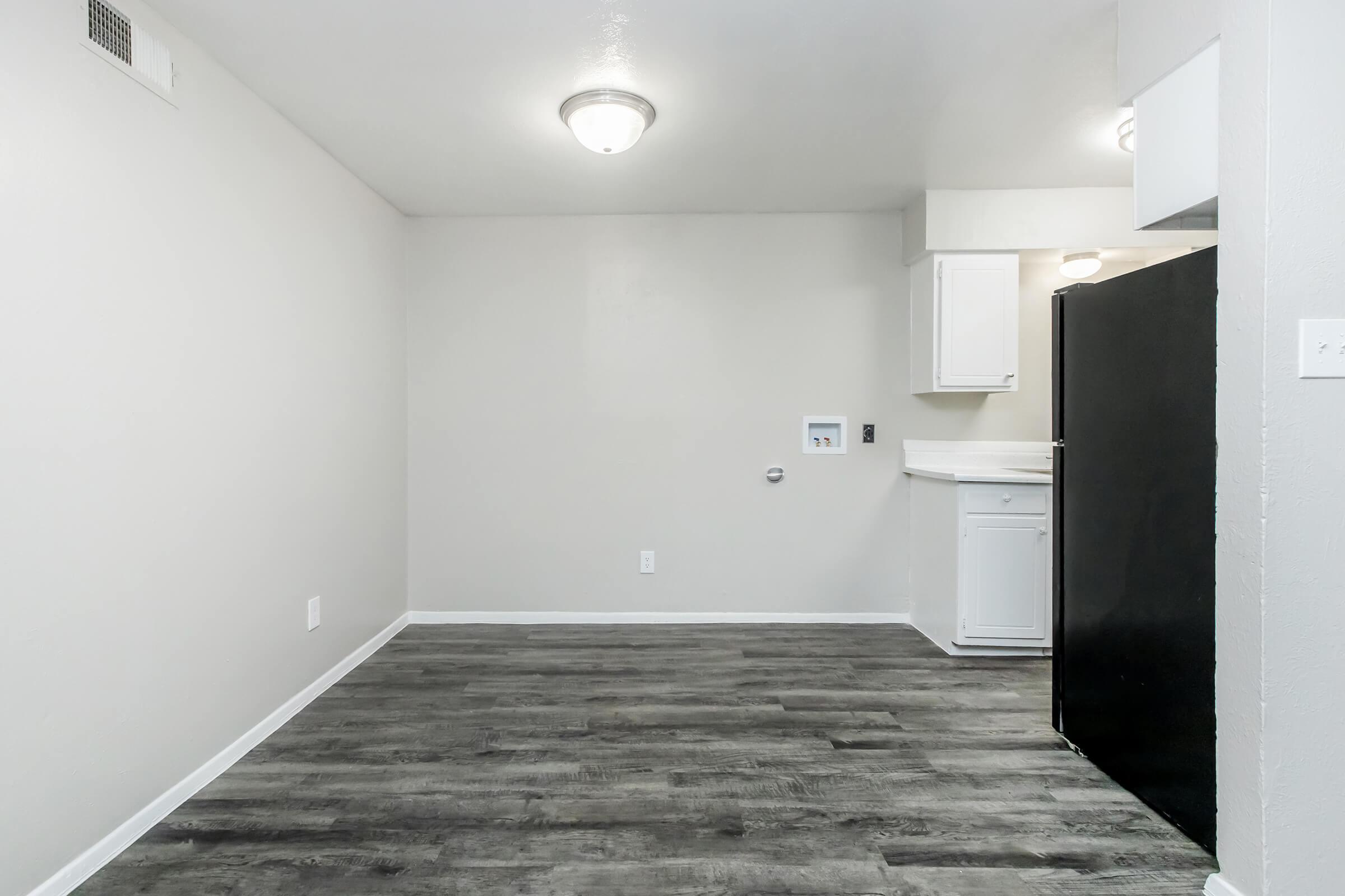A small, unfurnished room with light gray walls and a dark laminate floor. It features a ceiling light, a black refrigerator, and a small kitchen space with white cabinets. There is a single electrical outlet on the wall and a small area on the left where a window might typically be.