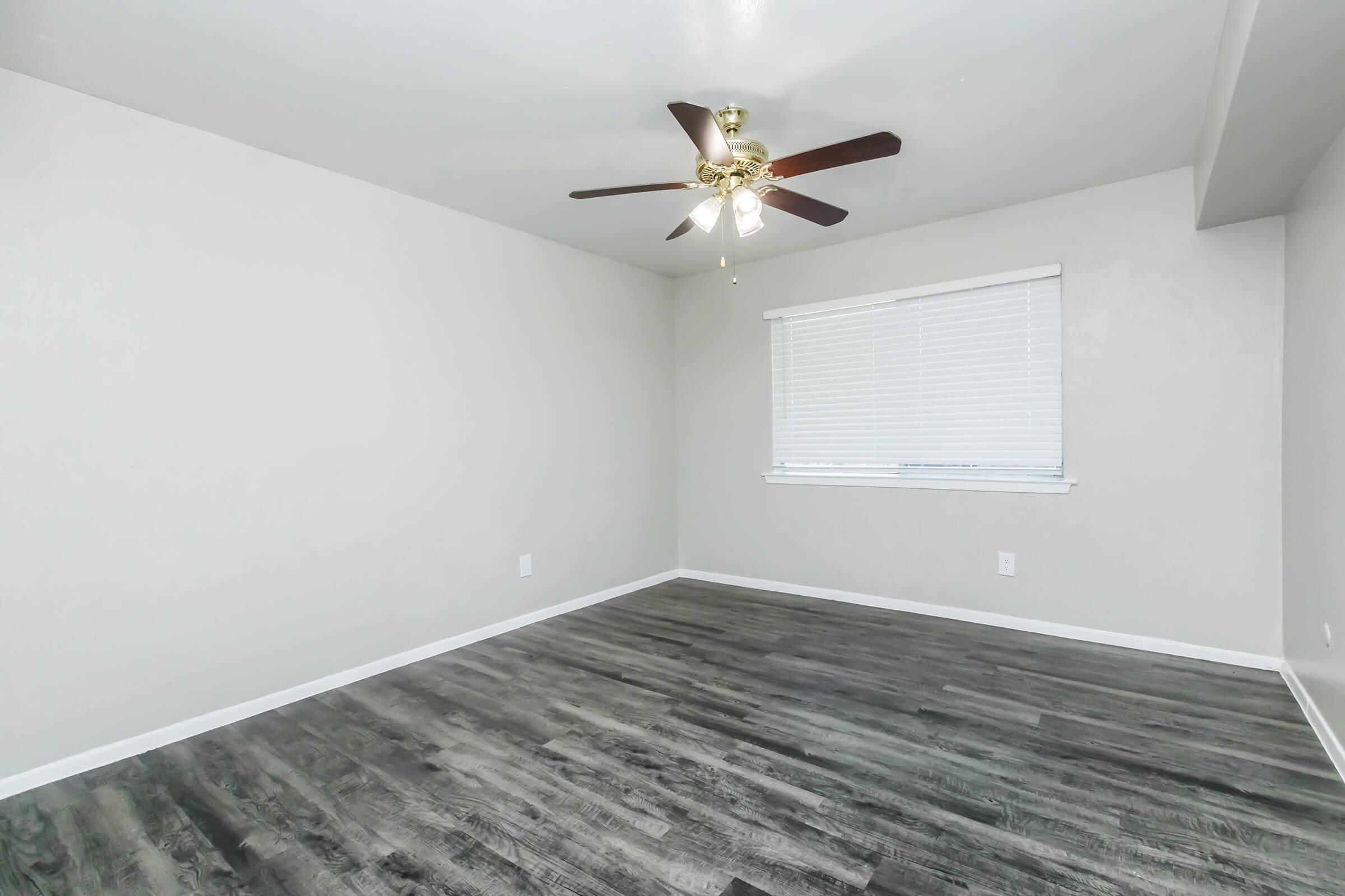 Empty room with light gray walls and a ceiling fan. The floor features dark wood-like planks. A window with white blinds allows natural light. The space is uncluttered, creating a minimalist aesthetic.