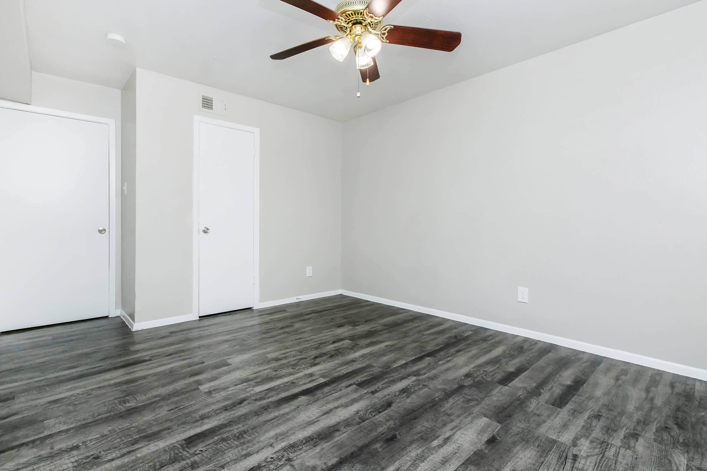 A spacious, empty room featuring light gray walls, a modern ceiling fan with wooden blades, and new dark wood-like laminate flooring. There is a closed white door on one side and a small closet door on the other, creating a bright and open atmosphere.