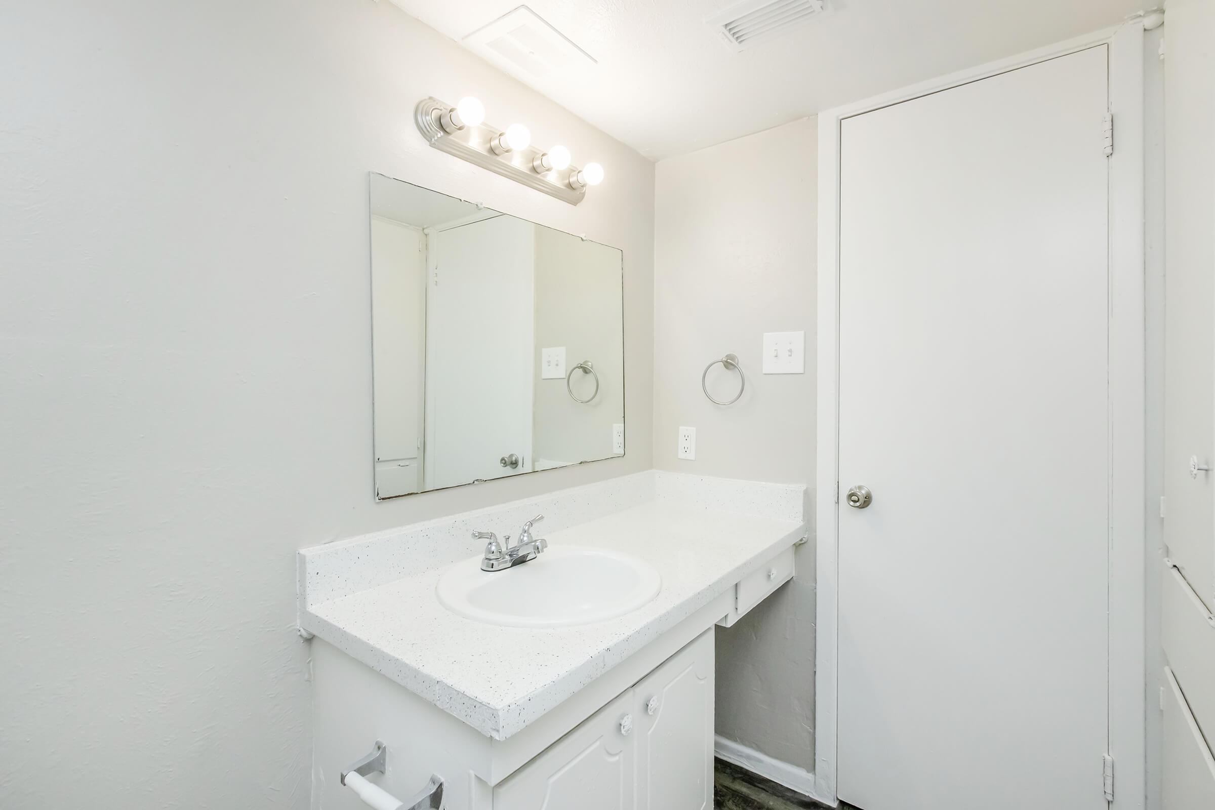 A clean and modern bathroom with light gray walls. Features include a white countertop with a circular sink, a large mirror above the sink, and a wall-mounted towel ring. The space also has a closed door and a small cabinet, all contributing to a bright and minimalist aesthetic.