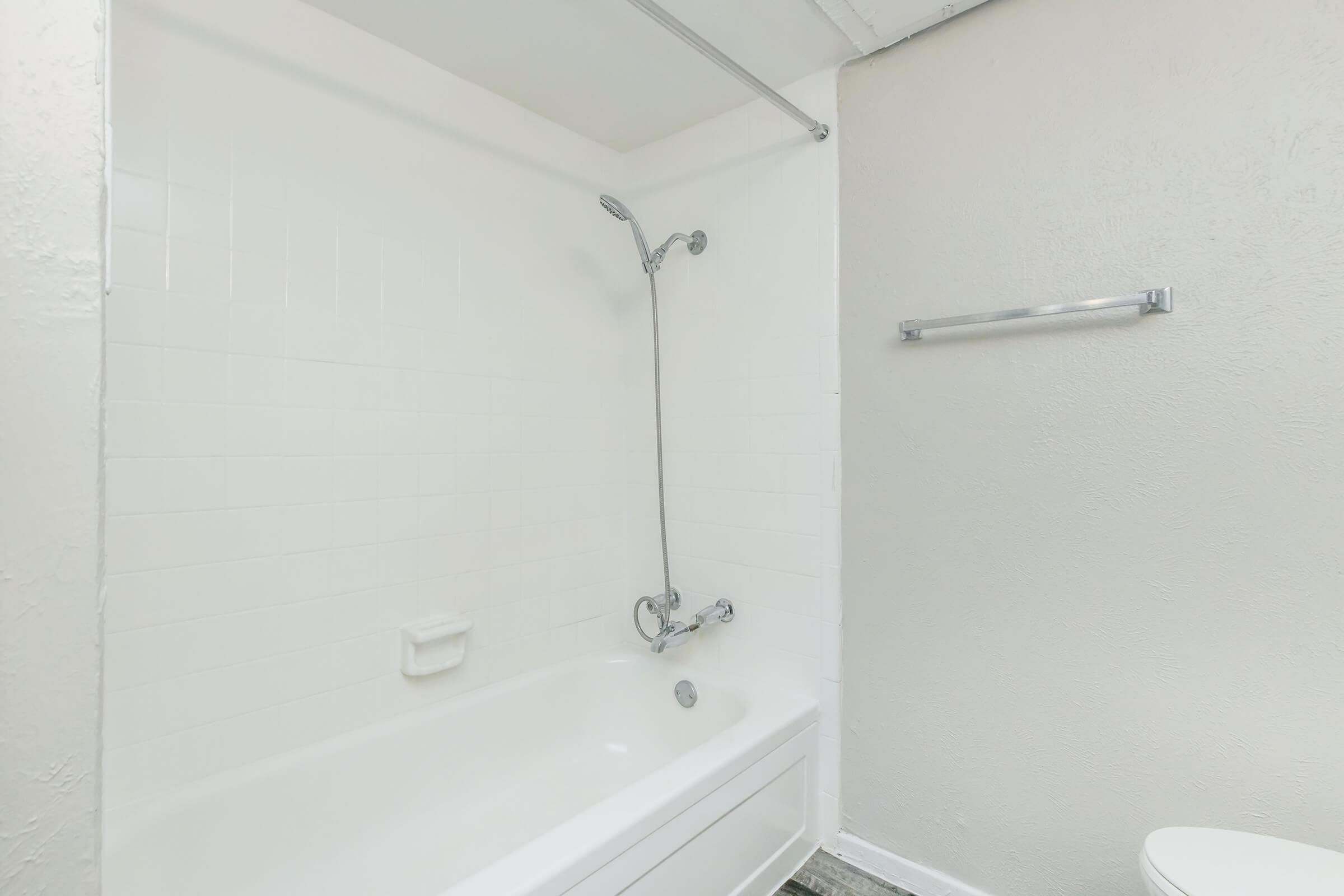 A clean, minimalist bathroom featuring a bathtub with a showerhead, white tiled walls, and a chrome towel bar. The space is bright with neutral tones, showcasing a modern design. The floor is partially visible, hinting at a simple and functional layout.