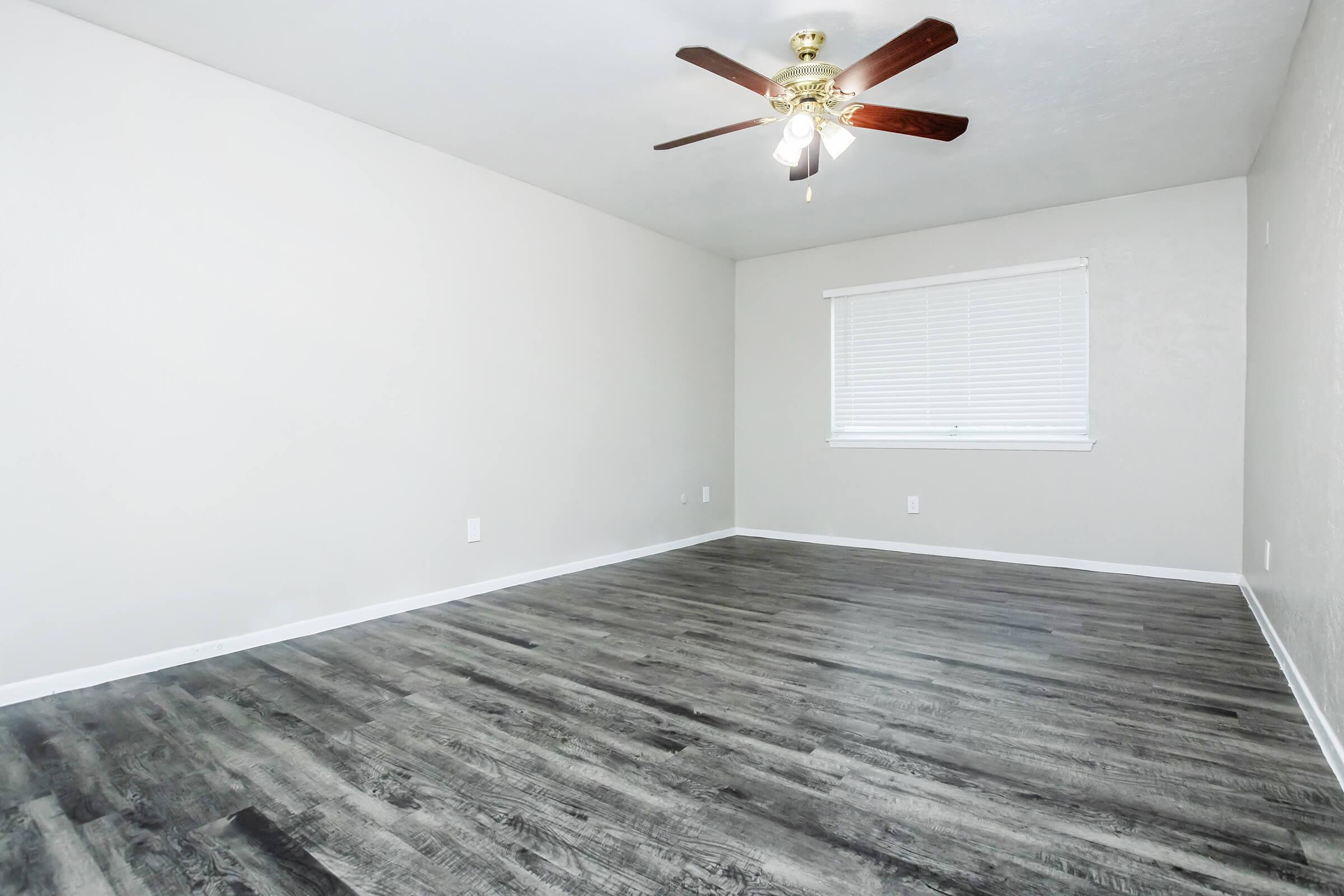 A spacious, empty room with light gray walls and dark wood-like flooring. A ceiling fan with a light fixture hangs from the ceiling. There is a window with white blinds allowing natural light to enter, and the overall ambiance appears bright and uncluttered, suitable for various furnishings.