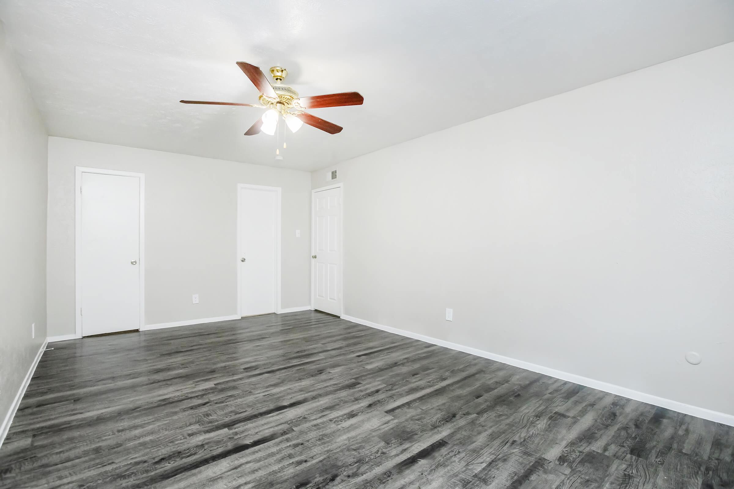 A spacious room with light gray walls and a ceiling fan. It features a wood-like floor and two closed white doors on the left side. The room is empty, creating a bright and airy atmosphere.