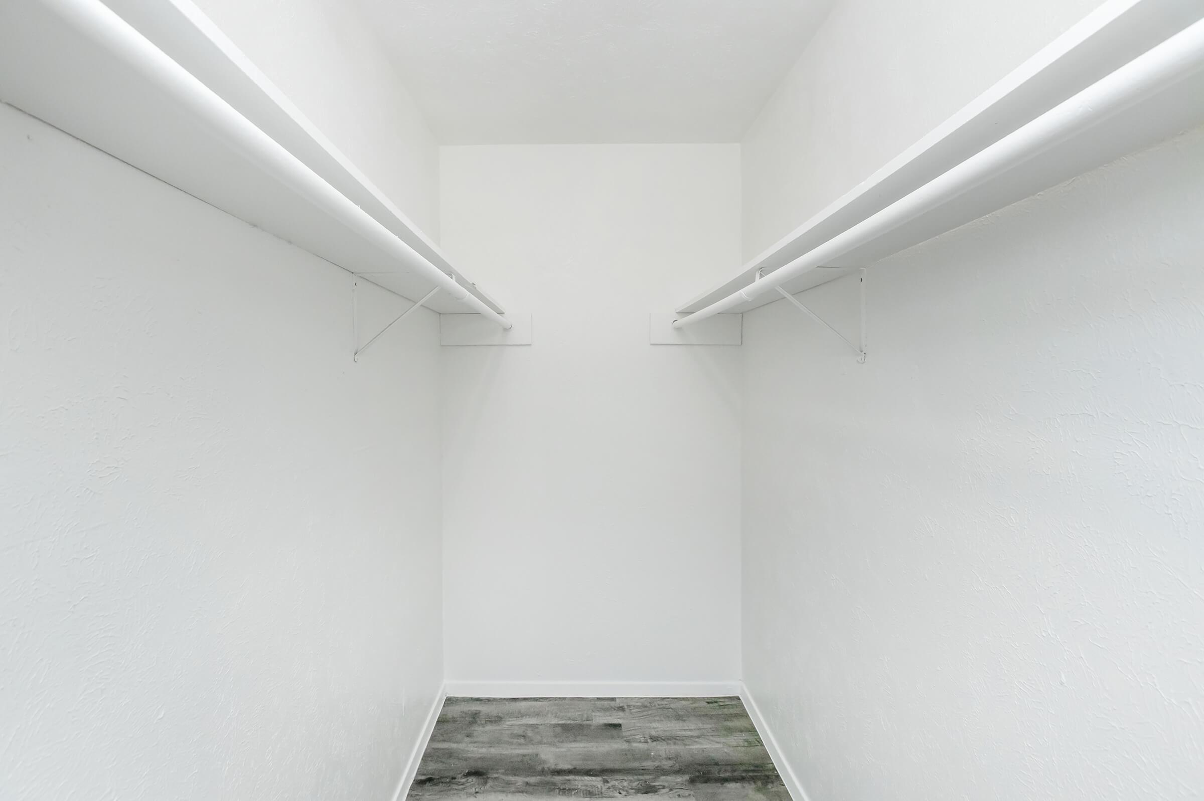 An empty walk-in closet with white walls and two hanging rods. The floor is a light-colored laminate. The space is minimal and uncluttered, with ample room for storage or organization.