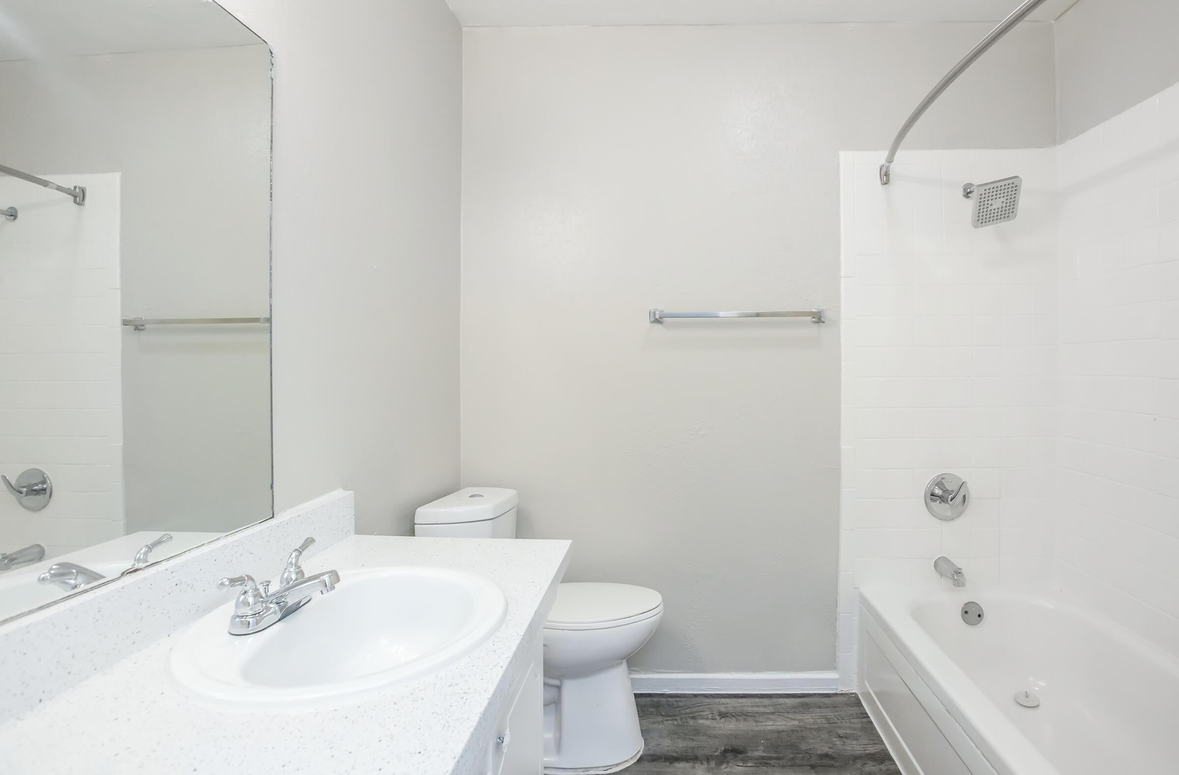 A modern bathroom featuring a white sink with a chrome faucet, a toilet, a bathtub with a shower curtain, and a large mirror above the sink. The walls are light-colored, and the floor is a wood-like finish, creating a clean and bright atmosphere.