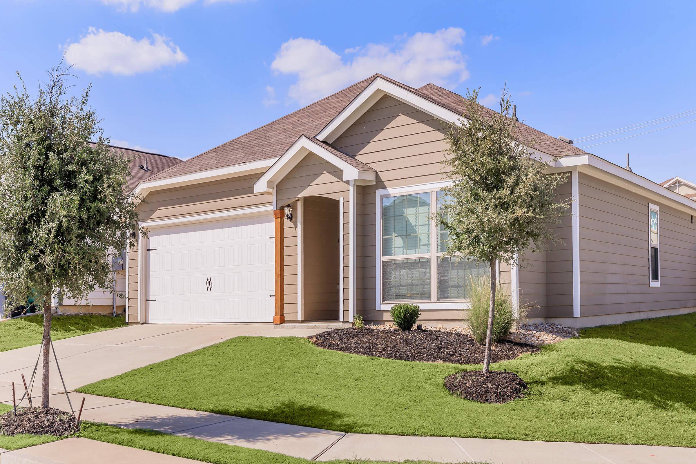 A modern single-story house with beige siding, a two-car garage, and a well-maintained front yard featuring green grass and small shrubs. The driveway leads up to the house, which has a welcoming entrance and large windows, set against a backdrop of blue sky and fluffy clouds.