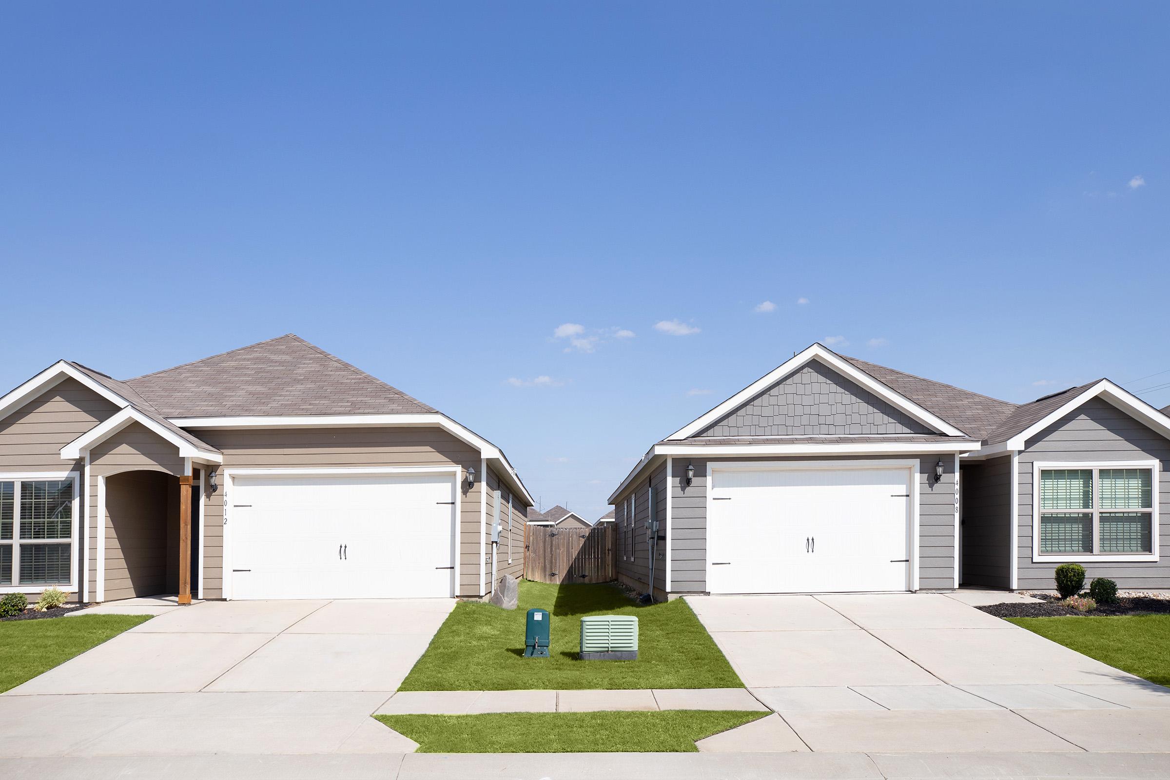 Two modern single-family homes with different exterior designs, one featuring light brown siding and the other gray. Both have garages, manicured lawns, and a clear blue sky in the background. Concrete driveways lead to the garages, enhancing a neighborhood look.