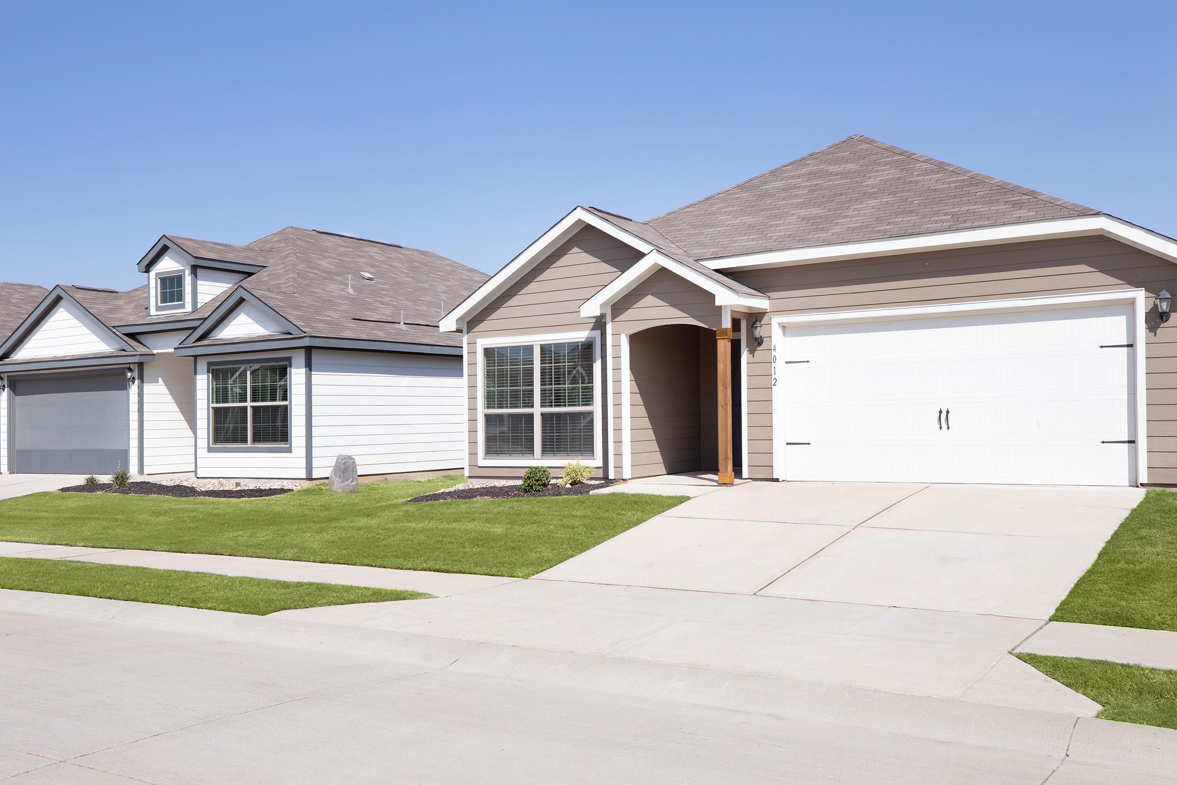 Two suburban homes side by side on a sunny day. The house on the right features a light brown exterior with a welcoming front porch, large windows, and a two-car garage. The left house is white with a darker roof and is slightly set back. Both homes have well-maintained lawns and clear blue skies above.
