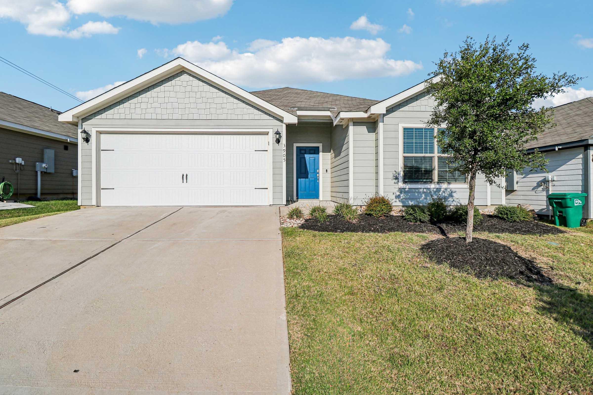 A modern single-story house with a light gray exterior, a blue front door, a two-car garage, and a well-manicured lawn. The driveway is paved, and there's a small tree and landscaped areas with shrubs around the front. The sky is bright with fluffy white clouds.