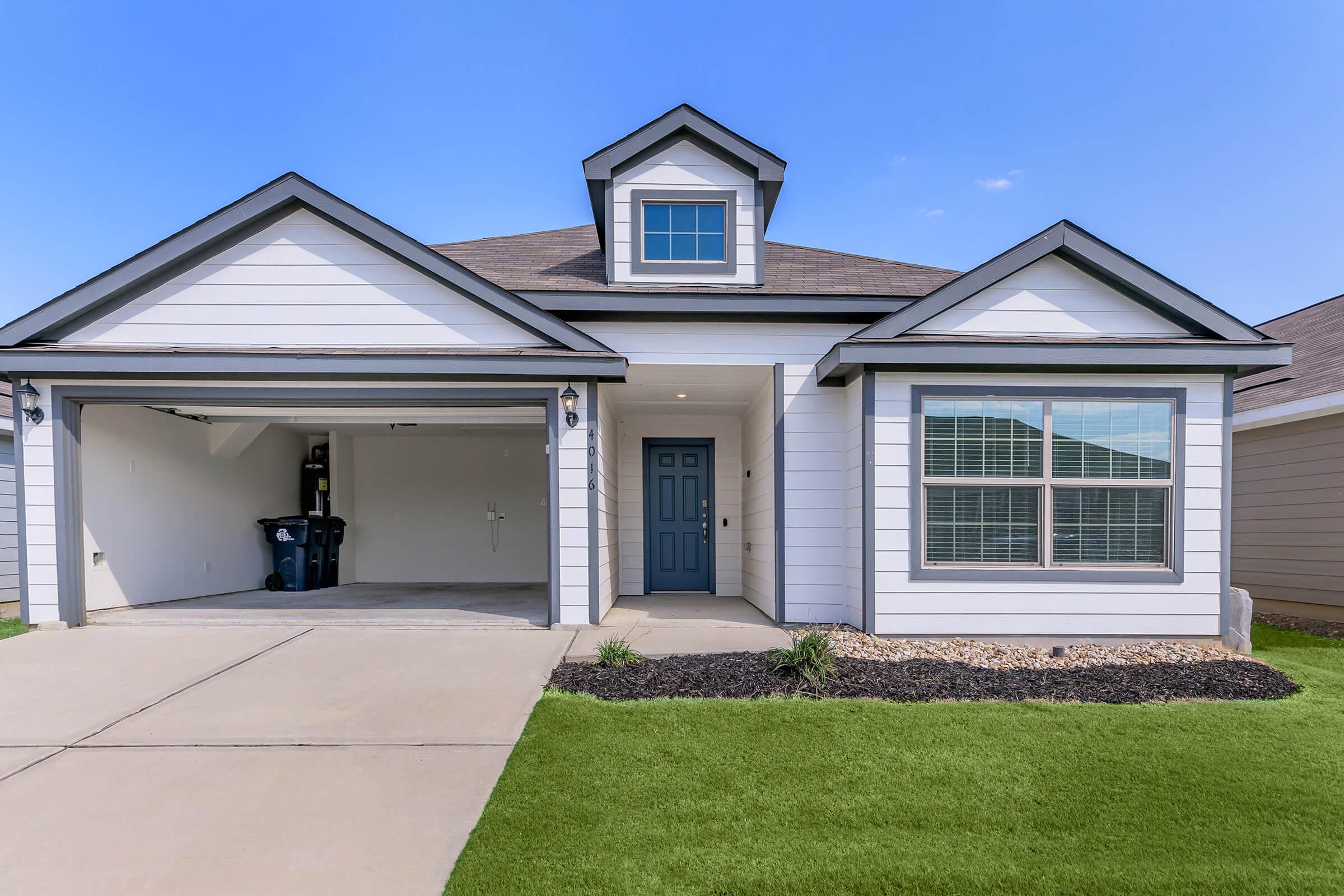A modern single-story house with a light gray exterior, a dark roof, and a small front porch. The garage door is slightly open, revealing an empty space inside. A large window on the right side features white trim and offers a view of the living area. The front yard has neatly manicured grass and decorative stones.