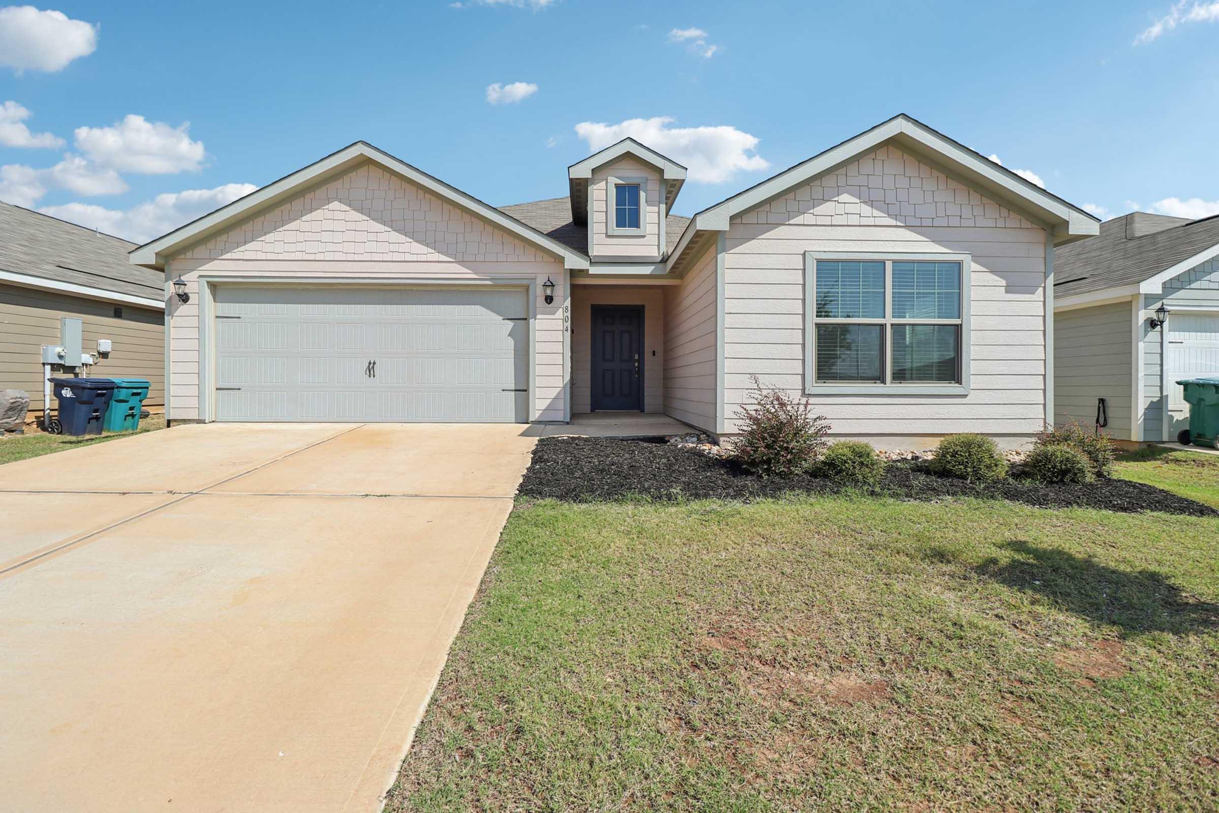 A single-story residential house with a gray exterior and a charming front porch. The driveway is paved, leading to a garage on the left. Lush green grass borders the driveway, and there are a few shrubs planted near the house. The sky is clear with a few fluffy clouds.