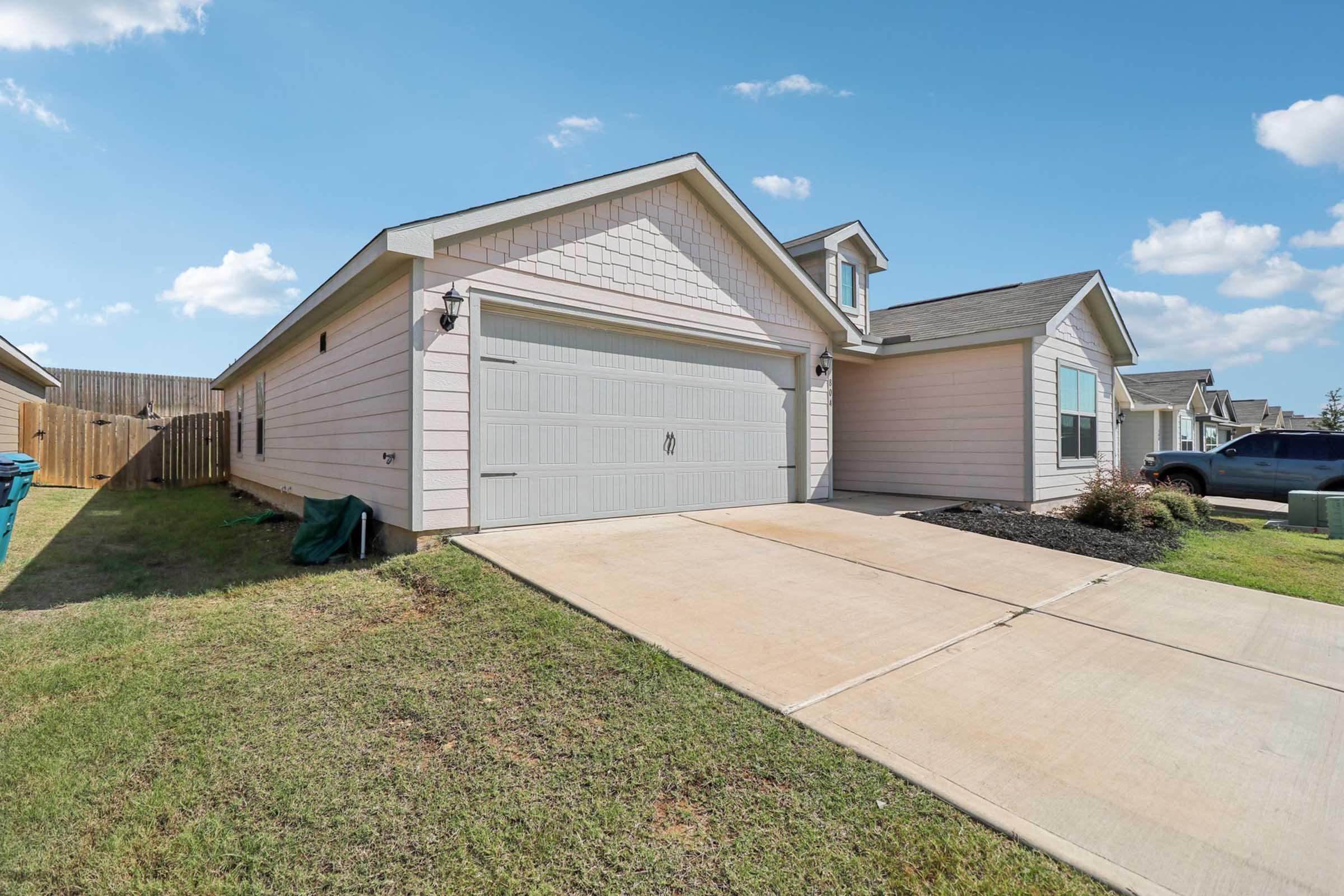 A modern single-story house with a light pink exterior, featuring a two-car garage and a paved driveway. The surrounding yard is well-maintained, with a wooden fence in the background and clear blue skies above. The home is set in a suburban neighborhood.