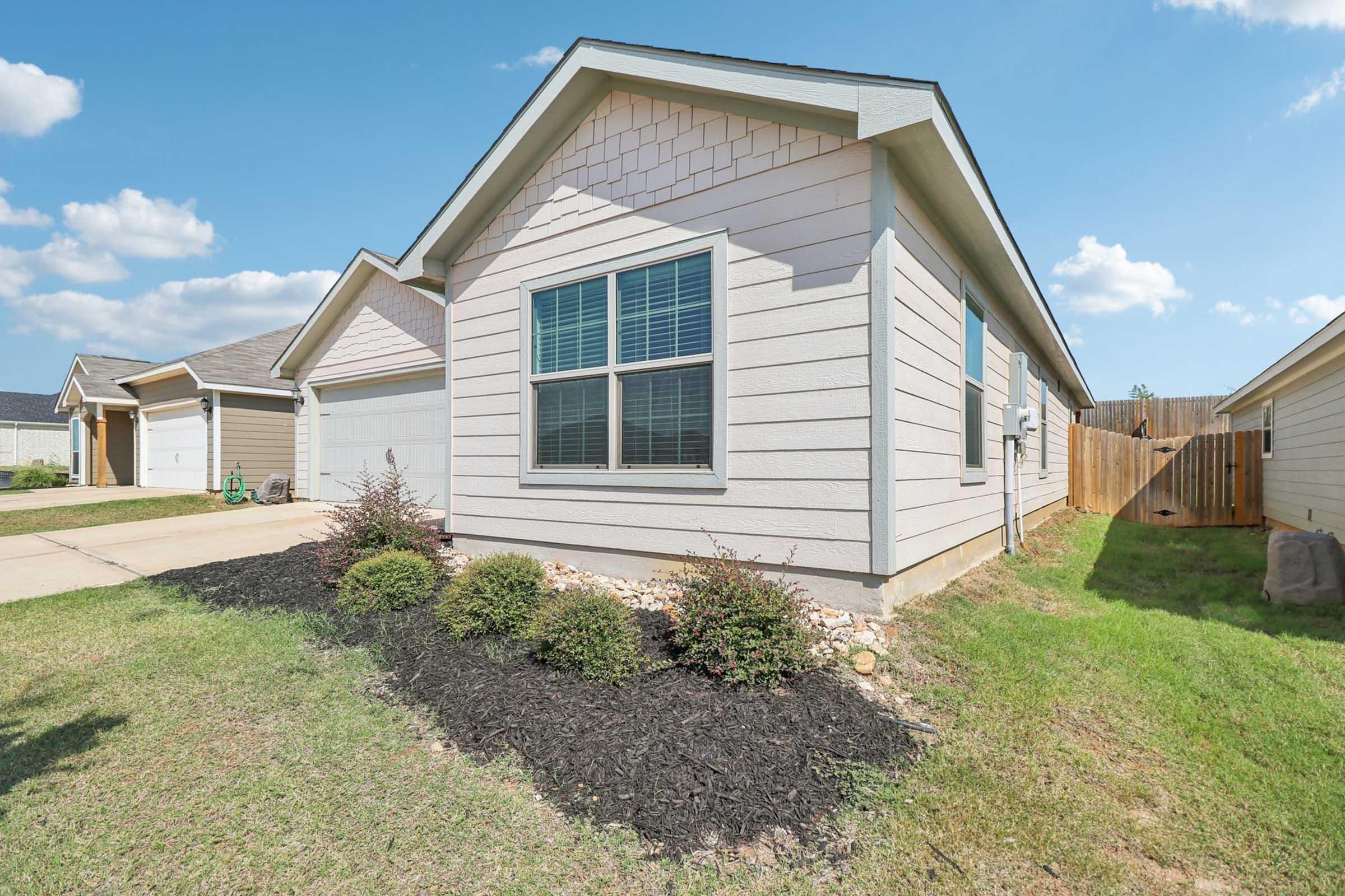 A modern single-story house with light-colored siding, featuring multiple windows and a neatly landscaped front yard with shrubs and mulch. The driveway leads to a garage, and a wooden fence is visible in the background against a clear blue sky.