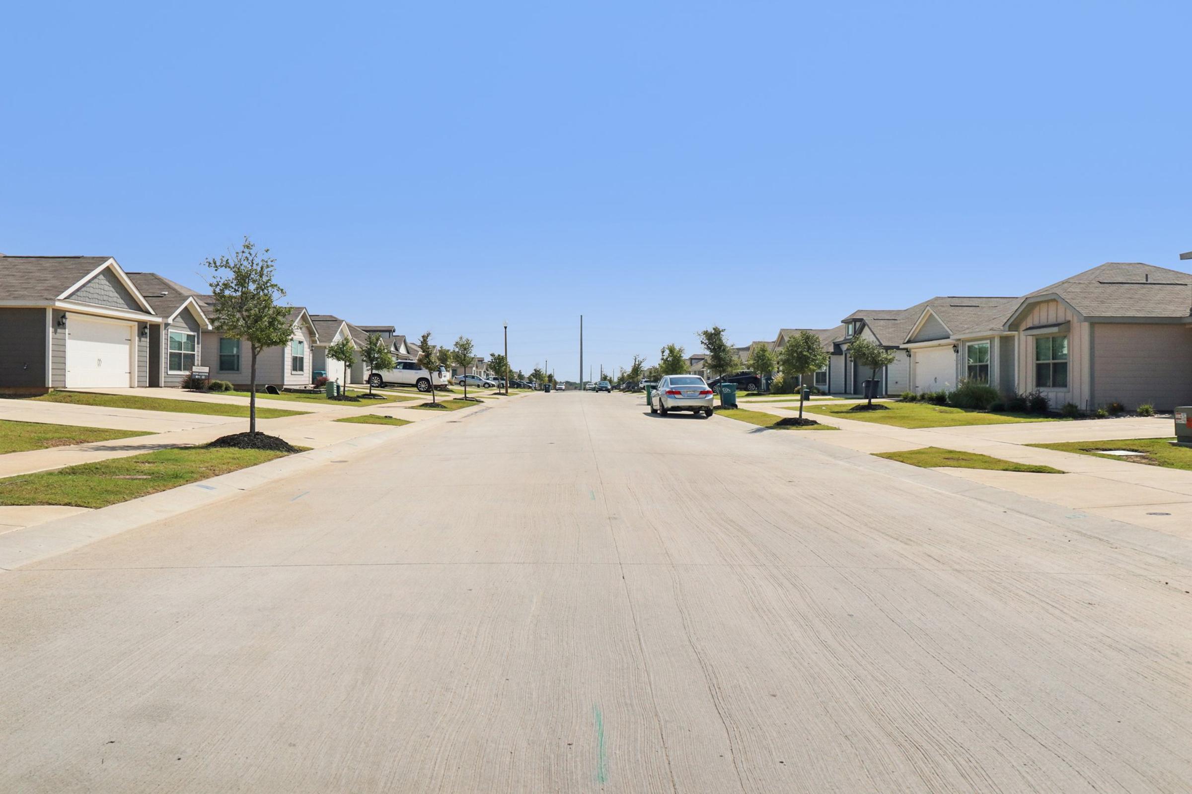 A quiet residential street lined with single-family homes under a clear blue sky. Grass and sidewalks are visible along both sides of the road, with a parked car on one side. The setting conveys a peaceful suburban atmosphere.