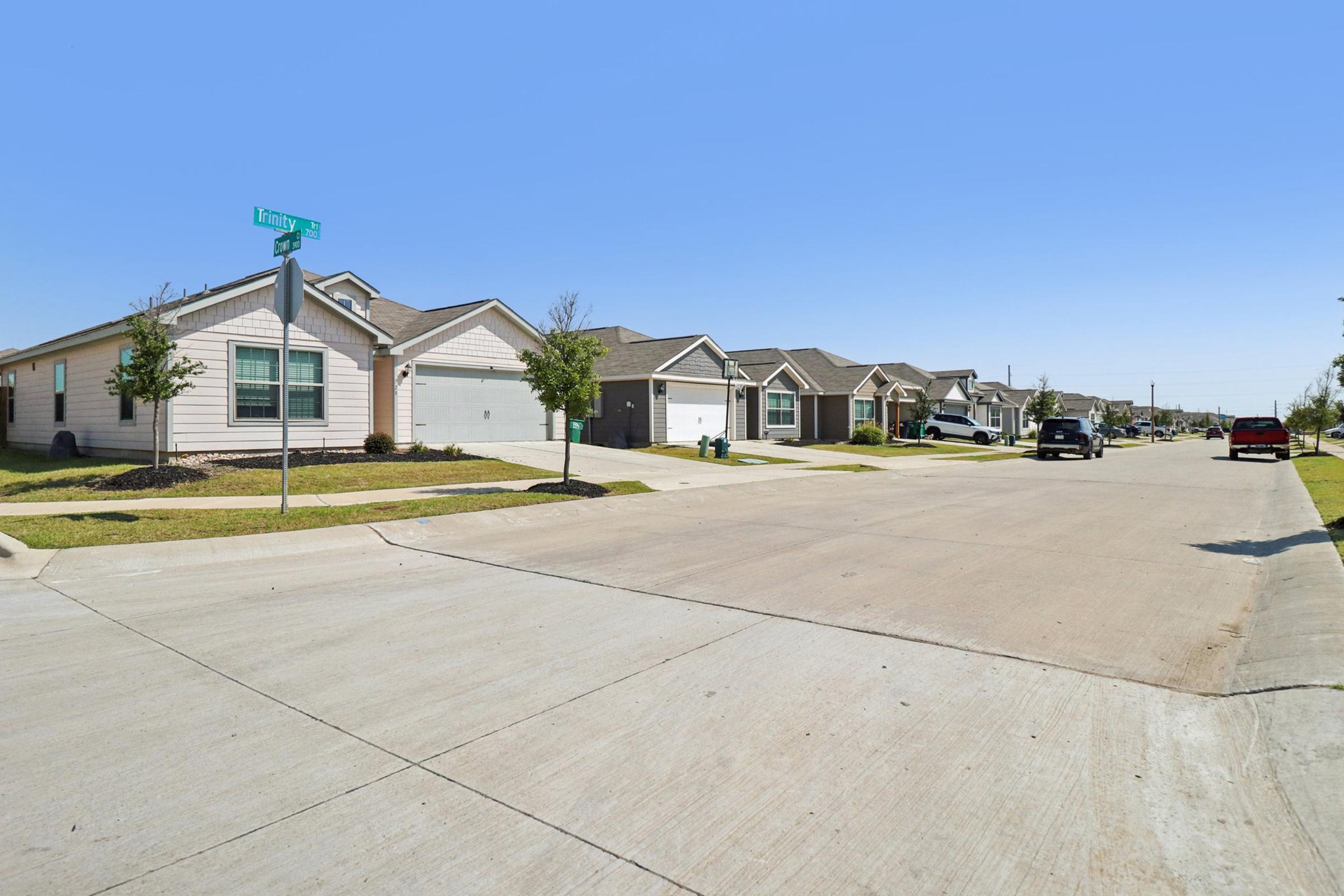 A quiet suburban street lined with single-family homes. The houses feature varied architectural styles, with well-maintained lawns and sidewalks. Clear skies above indicate a sunny day, and a few cars are parked along the street. A street sign is visible at the corner, labeling the intersection.