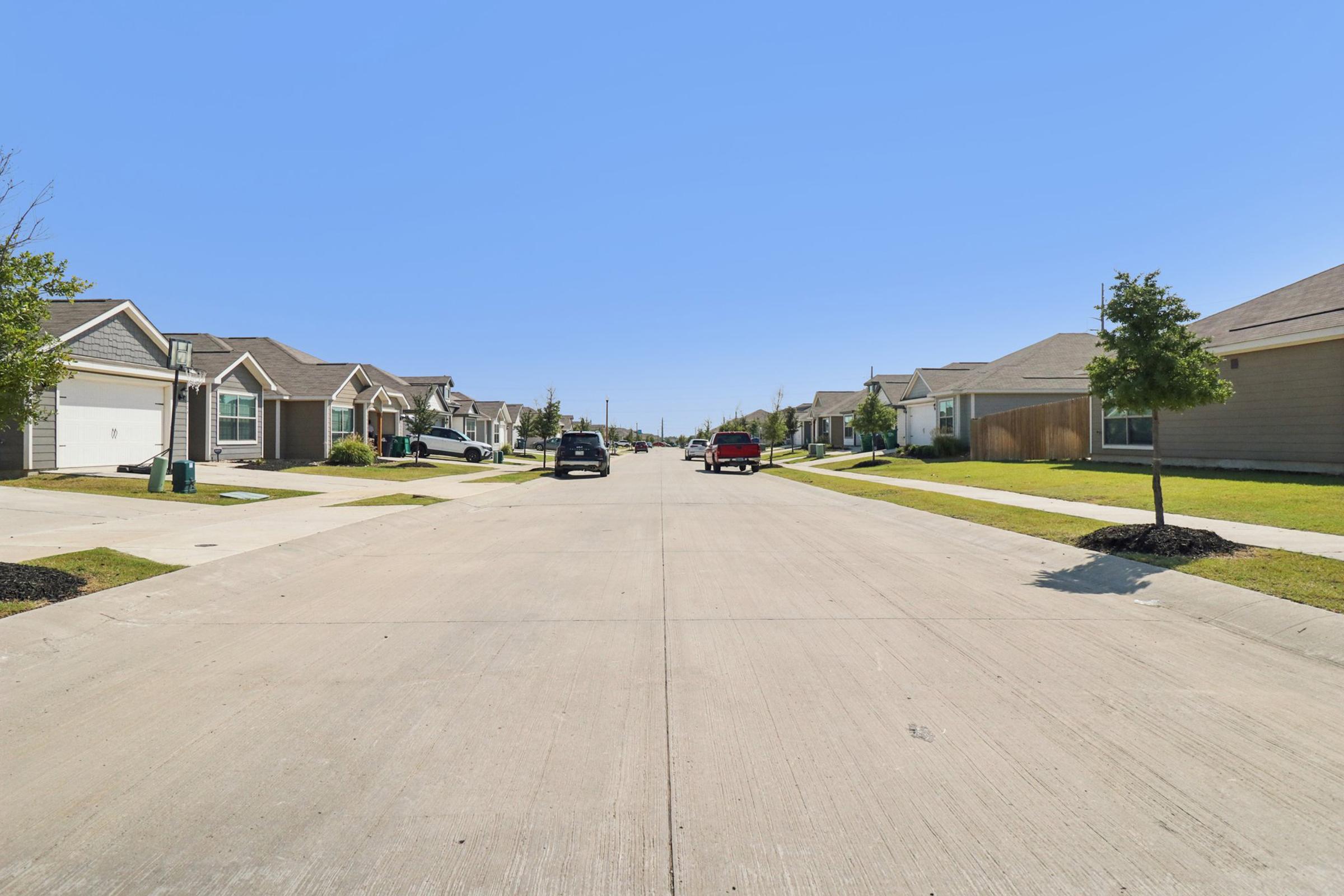 A quiet residential street lined with single-family homes. The road is paved and leads into the distance, with a clear blue sky overhead. Cars are parked on the sides, and well-maintained lawns with small trees are visible, contributing to a peaceful suburban atmosphere.