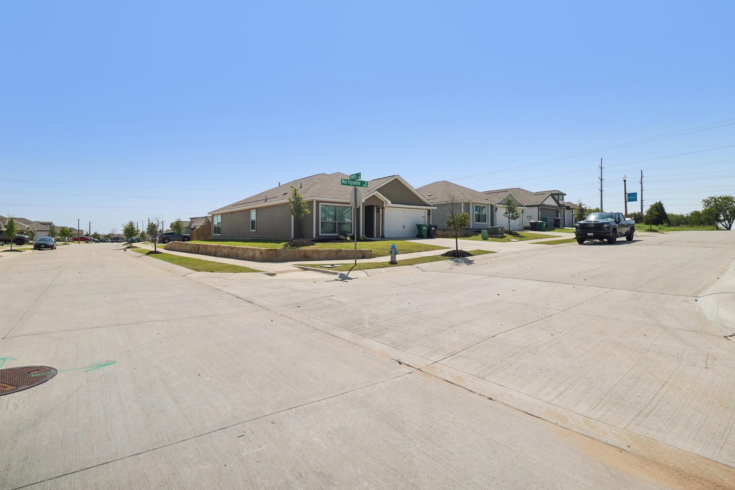 A street intersection in a suburban neighborhood featuring several houses. The view includes well-maintained lawns, pavement, and street signs. A black vehicle is parked at the corner, and the sky is clear and blue. The scene conveys a peaceful, residential atmosphere.