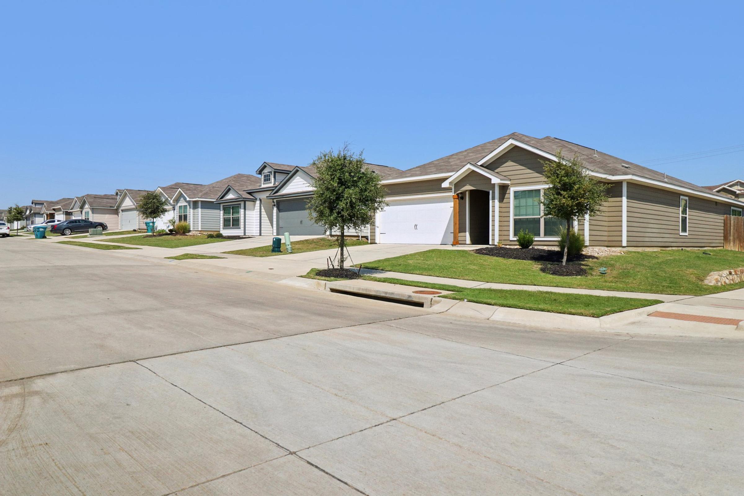 Residential neighborhood featuring several single-family homes with well-maintained lawns, driveways, and sidewalks under a clear blue sky. The street appears quiet, with a few trees and parked cars visible. Decoratively landscaped yards enhance the suburban atmosphere.