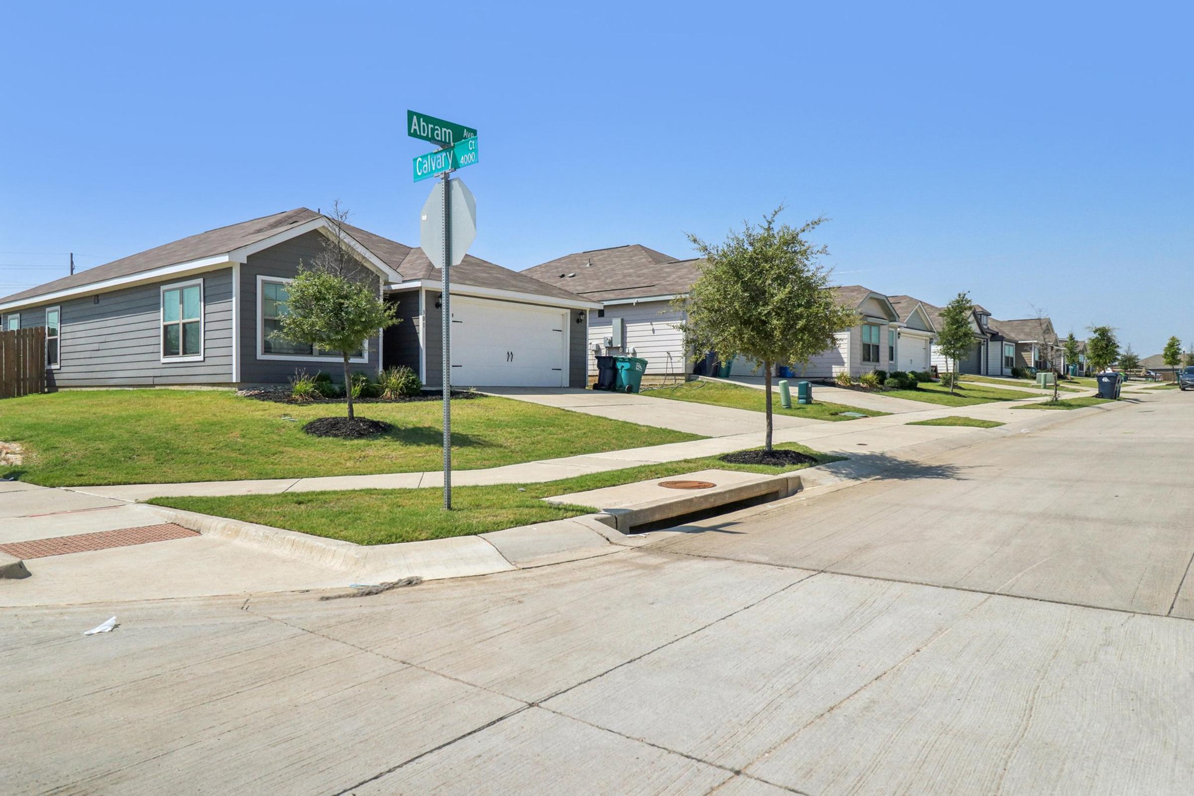 A quiet residential street featuring several houses with manicured lawns, driveways, and mailboxes. A street sign at the corner indicates "Abram St" and "Cato Ln." The sky is clear and blue, enhancing the suburban atmosphere.