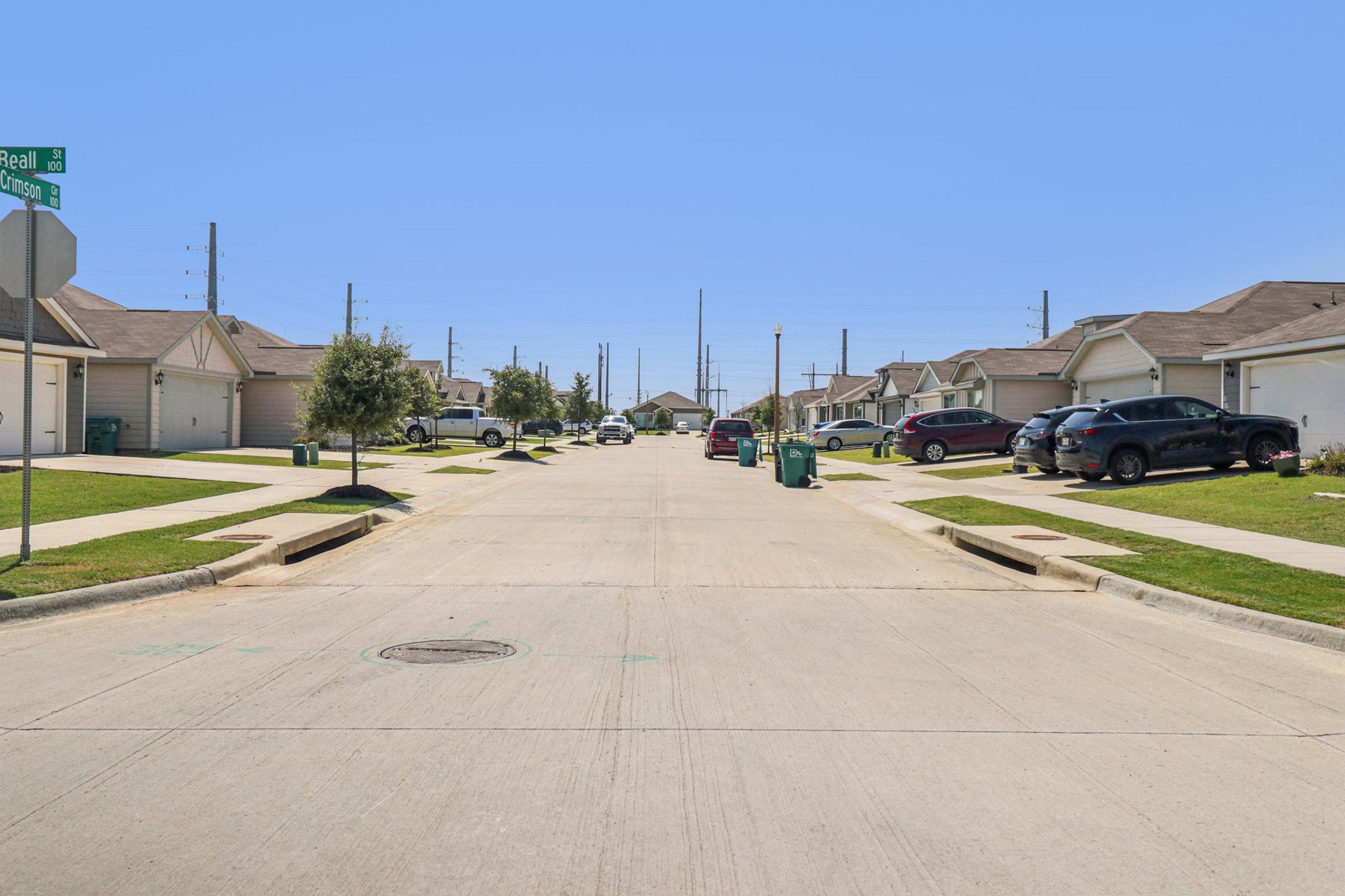 A residential street featuring single-family homes on either side. Vehicles are parked along the curbs, and green trash bins are placed near the sidewalk. Utility poles line the street, which has a clear blue sky overhead, creating a bright and sunny atmosphere.