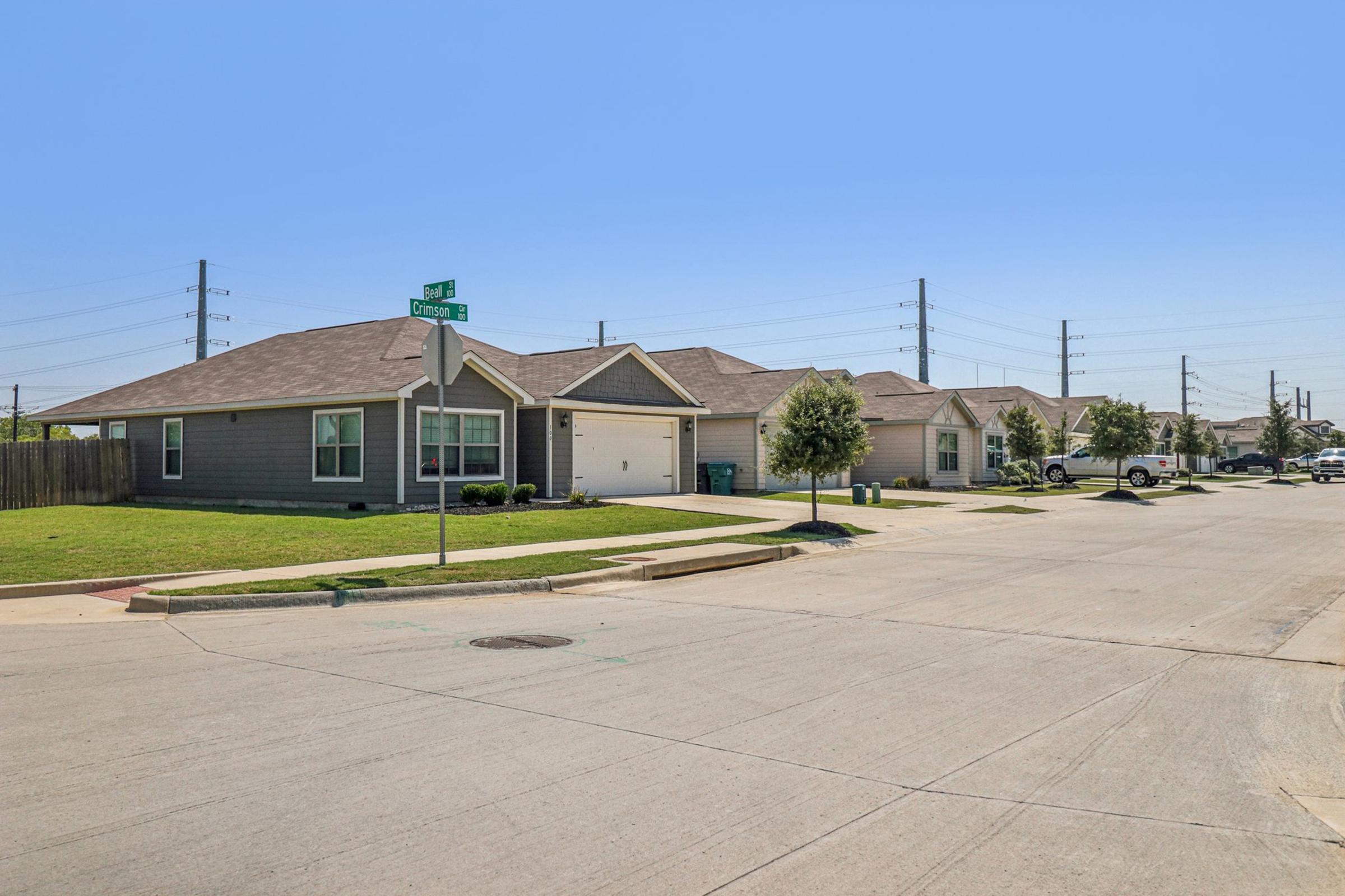 A quiet residential street featuring several single-story houses with gray siding. The homes have well-maintained lawns, a few small trees, and driveways. Power lines run overhead, and the street is paved with no visible pedestrians or cars. A street sign is partially visible in the corner.