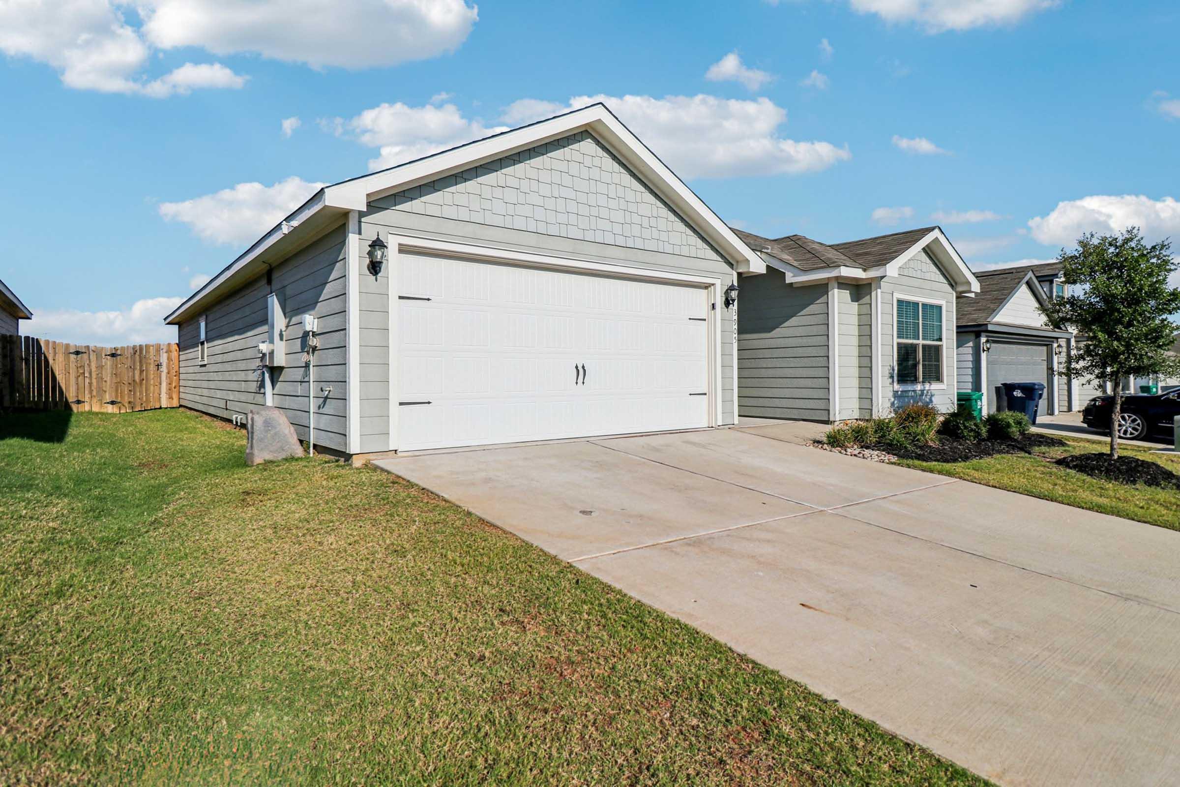 A freshly painted single-family home featuring a two-car garage with white doors. The driveway is made of concrete, and the front yard is green and well-maintained. Decorative shrubs line the house, and a wooden fence is visible in the background under a bright blue sky with fluffy clouds.