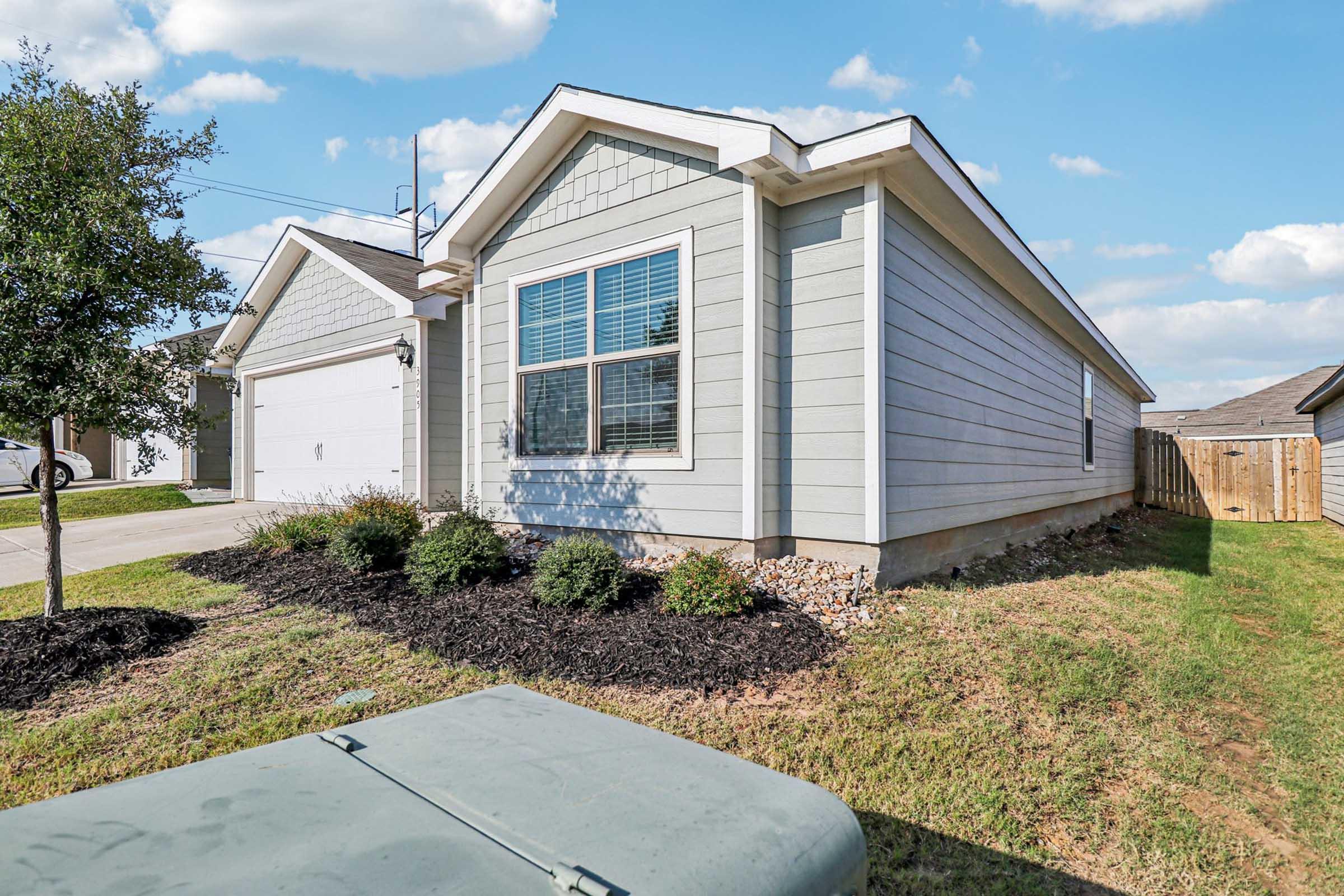 A modern single-family home with light gray siding and a neatly landscaped yard featuring shrubs and fresh mulch. The house has a two-car garage and a large window on the front facade, surrounded by green grass. In the foreground, there's a utility box beside the house. The sky is bright with a few clouds.