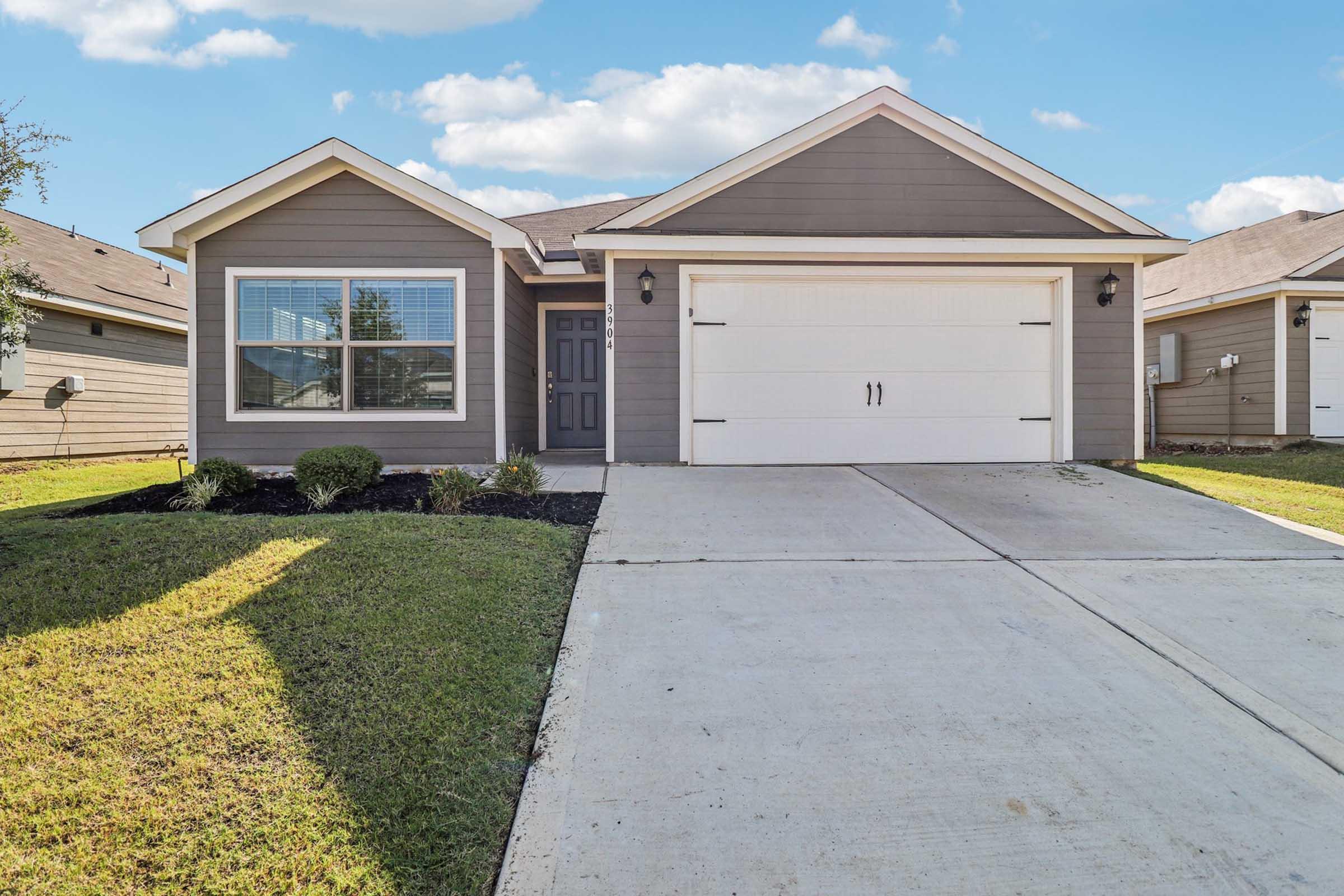 A modern single-story house with a light grey exterior, featuring a two-car garage, neatly landscaped front yard, and a concrete driveway. Windows are framed in white, and the door is dark with a decorative light fixture beside it. The sky is blue with a few clouds.