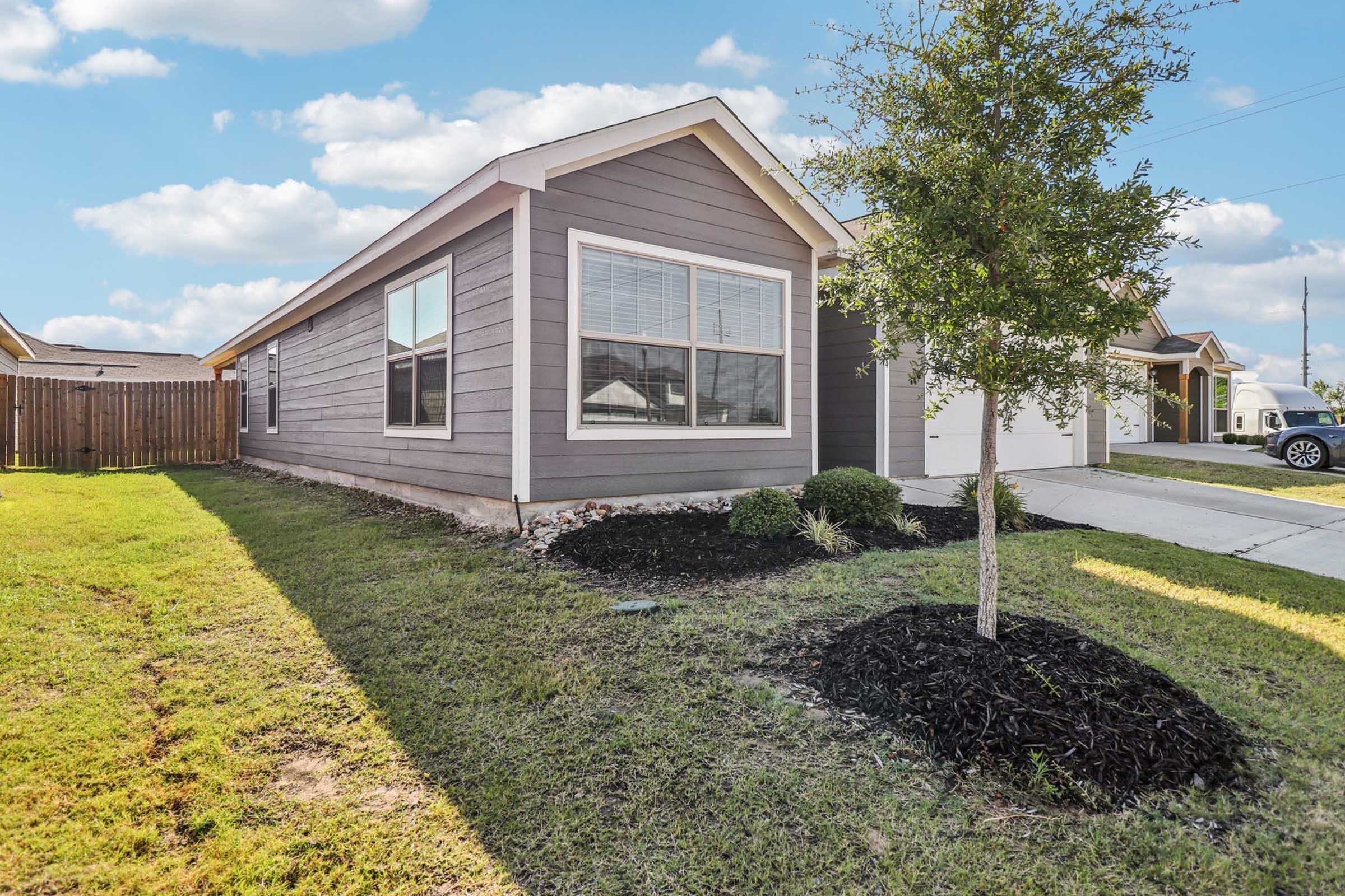 A modern single-story house with gray siding, featuring large windows and a neatly landscaped front yard. The lawn is well-maintained, with a small tree and black mulch around shrubs. The driveway leads to a garage, and there is a wooden fence in the background, indicating a suburban setting.