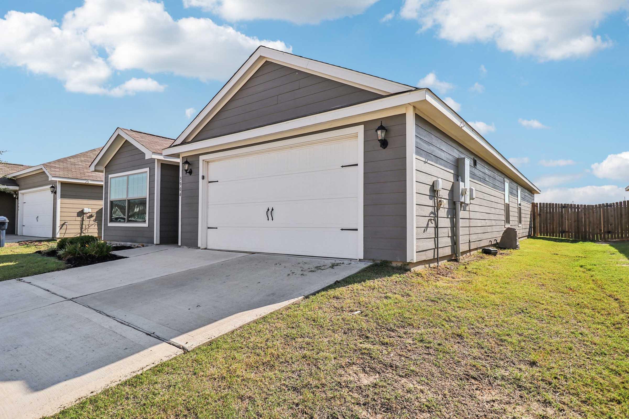 A gray house with a white garage door and a concrete driveway, set against a clear blue sky with fluffy white clouds. The front yard is well-maintained with grass. A neighboring house is visible to the left, and there is a wooden fence in the background.