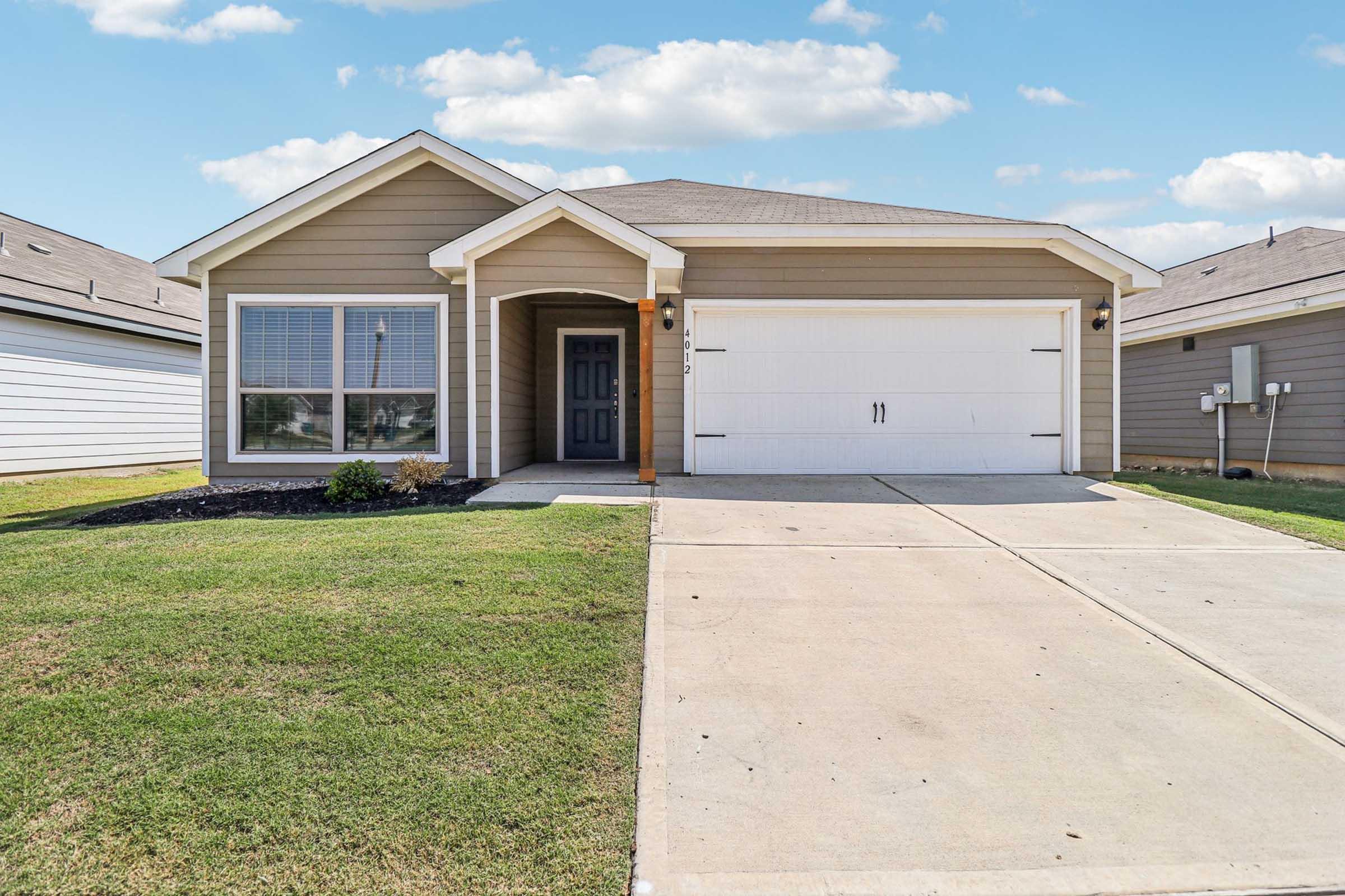 A modern single-story house with a light brown exterior, a two-car garage, and a neatly landscaped front yard. The house features a front porch with a blue door and large windows. The driveway is concrete, leading up to the garage, and the sky is partly cloudy.