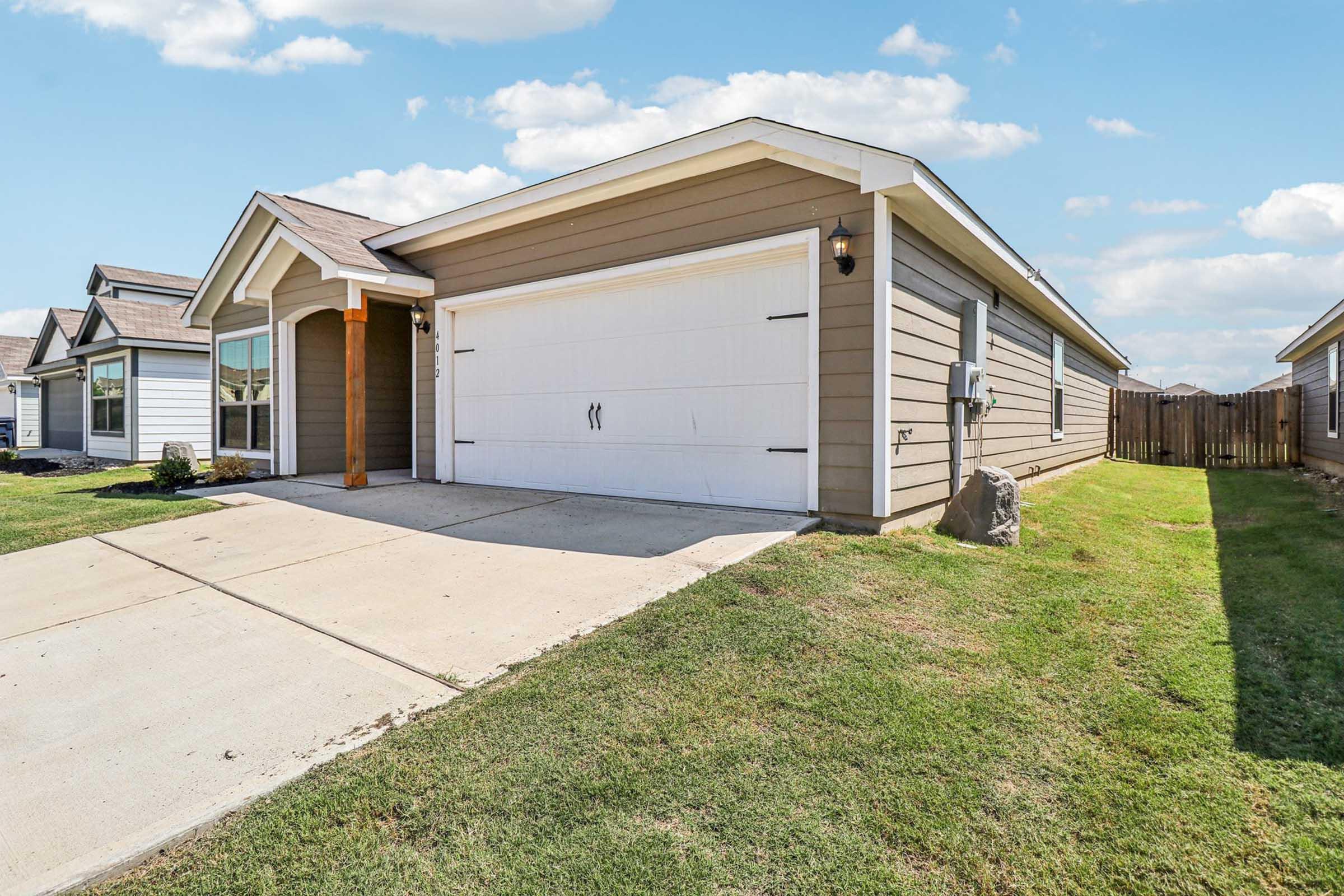 A well-maintained single-family home with a neatly landscaped front yard. The house features light brown siding, a spacious driveway, and a white garage door. Nearby are additional homes with similar architecture, set against a clear blue sky with a few clouds.