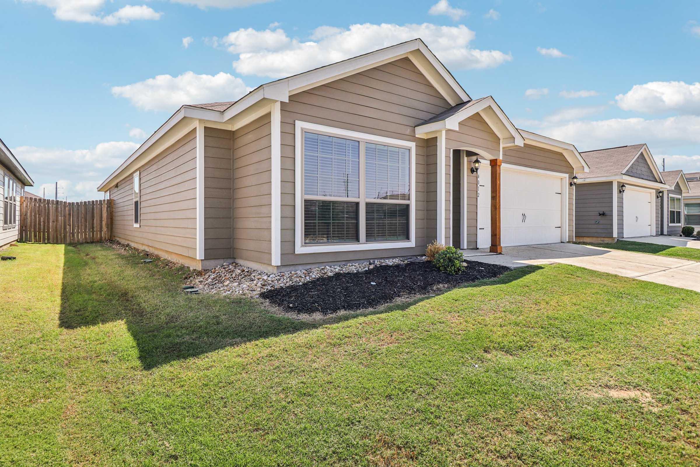 Single-story house with light brown siding and white trim. Features a well-maintained front yard with green grass and small shrubs. There are large windows on the front, and the driveway leads to an attached garage. The property is surrounded by a wooden fence, with clear blue skies in the background.
