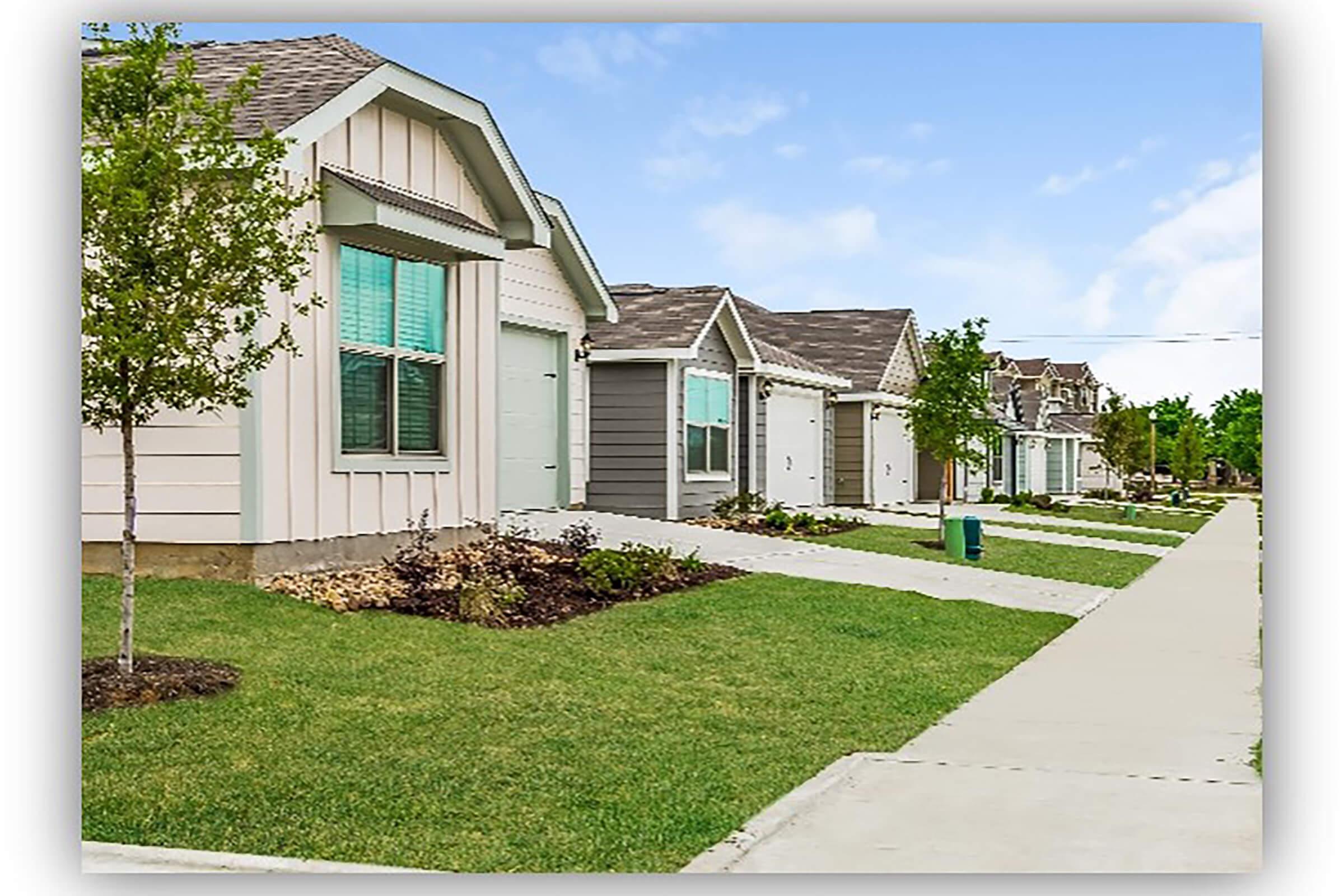 A row of modern, single-story homes with varying colors, set on a well-maintained street. Each house features a small front yard with grass and landscaping, and sidewalks leading to the entrances. The scene is bright with clear blue skies in the background.