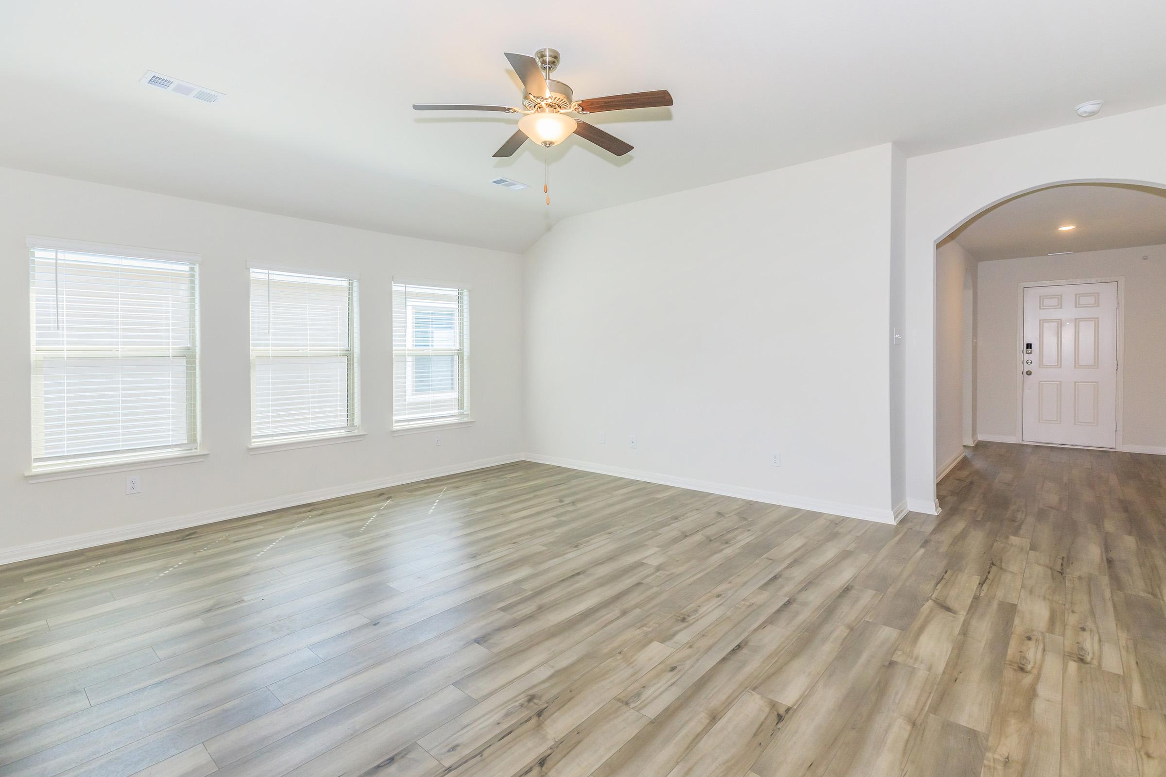 Spacious, empty living room with large windows allowing natural light, light-colored walls, and wooden flooring. A ceiling fan hangs above, and an entrance hallway with a front door is visible to the right. The room has a bright and airy feel.