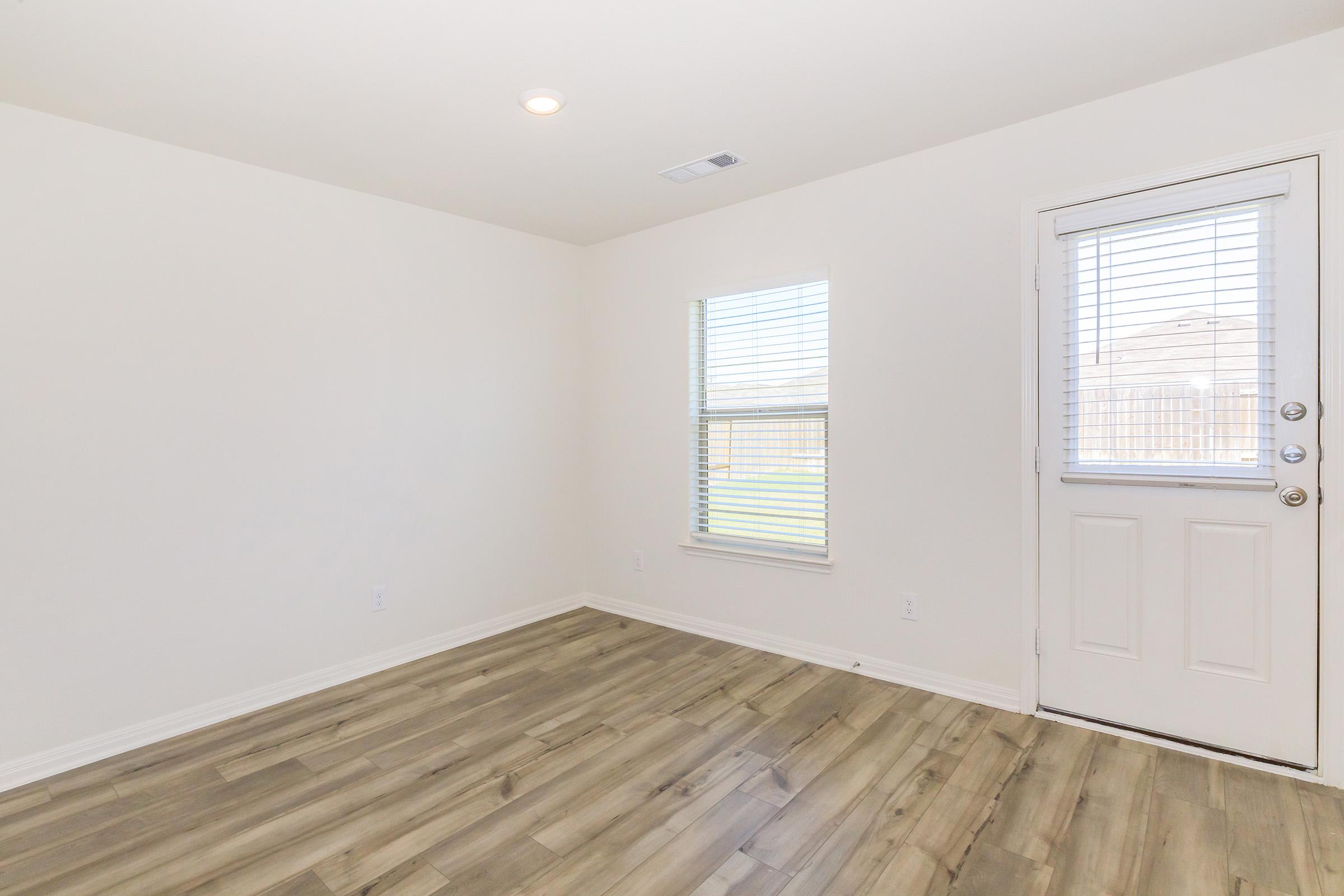 A bare room with light-colored walls and a wooden floor. It features a window with blinds on the left side and a door on the right that opens to the outside. The space is well-lit with natural light, creating an airy atmosphere.