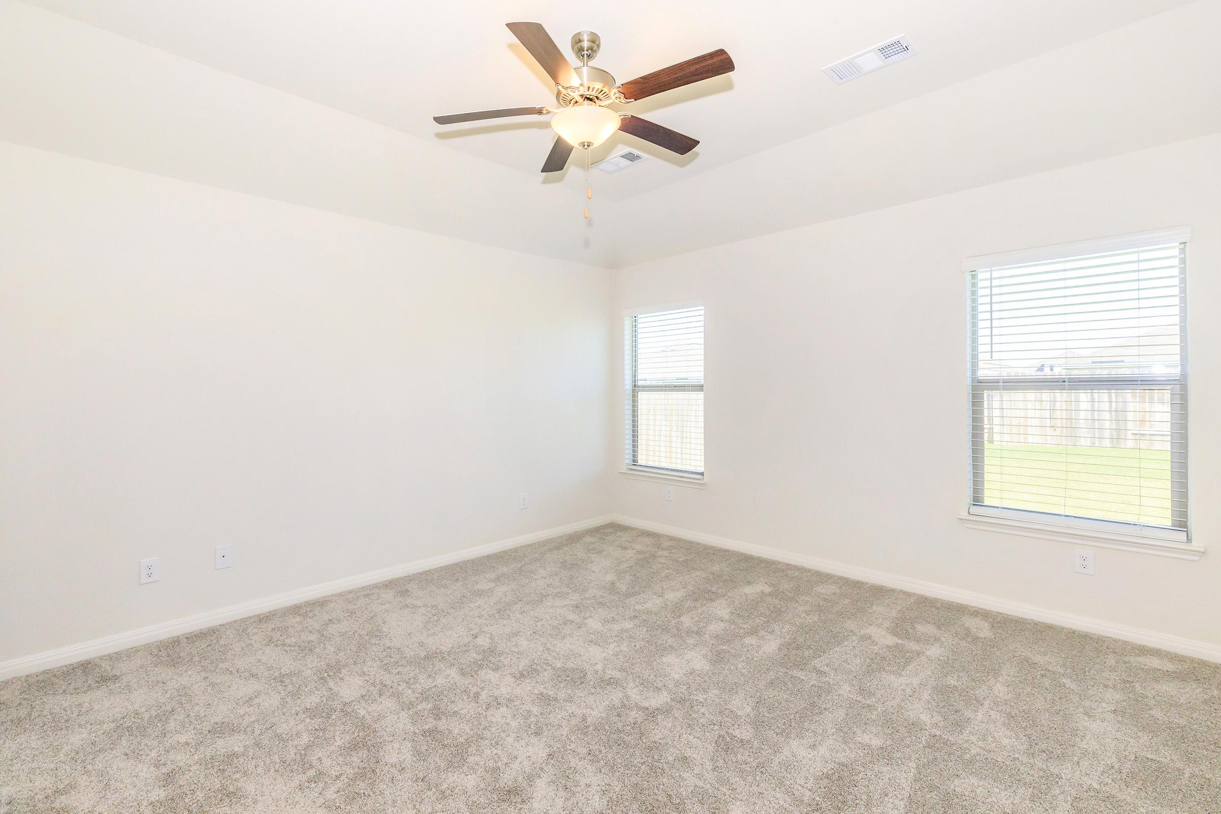 Empty room with light gray carpet, two windows with blinds, and a ceiling fan. The walls are painted light beige, giving a bright, airy feel. The space is unfurnished, providing a blank canvas for interior design.